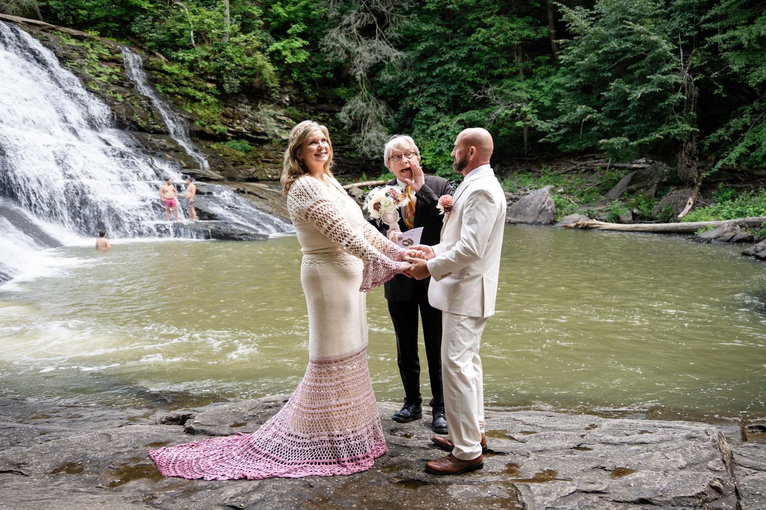 Couple exchanges vows before a waterfall with an officiant, bride in lace gown, groom in cream suit, outdoor wedding.