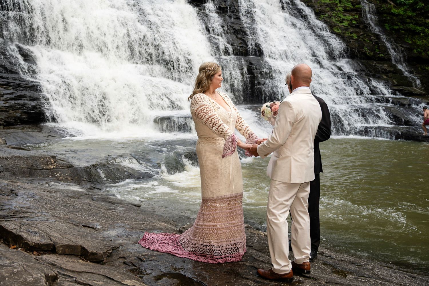 Couple exchanging vows in front of a stunning waterfall, bride in lace gown holding bouquet, groom in cream suit.