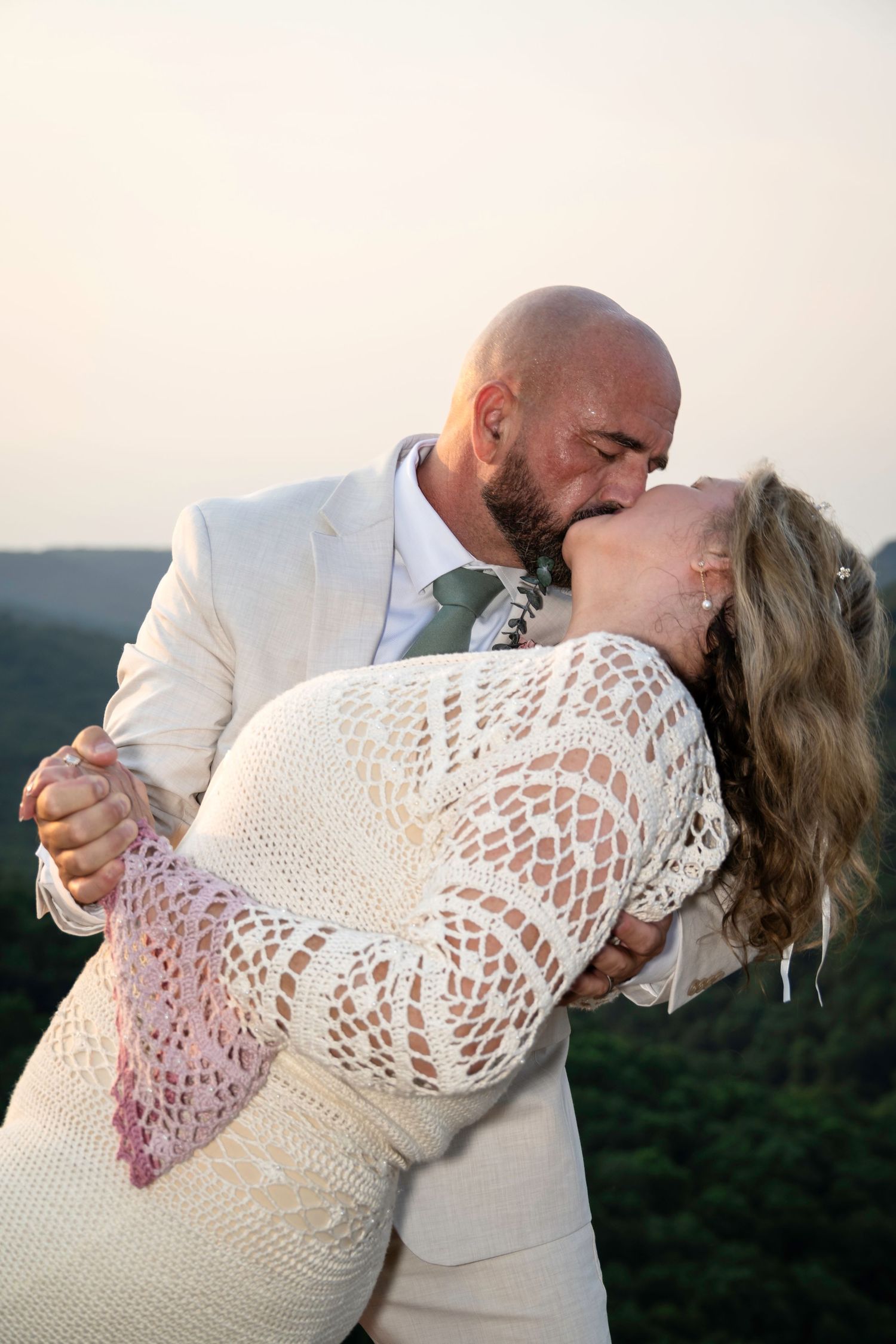 Bride and groom share a romantic dip kiss outdoors at sunset, wearing elegant wedding attire with green hills in background.
