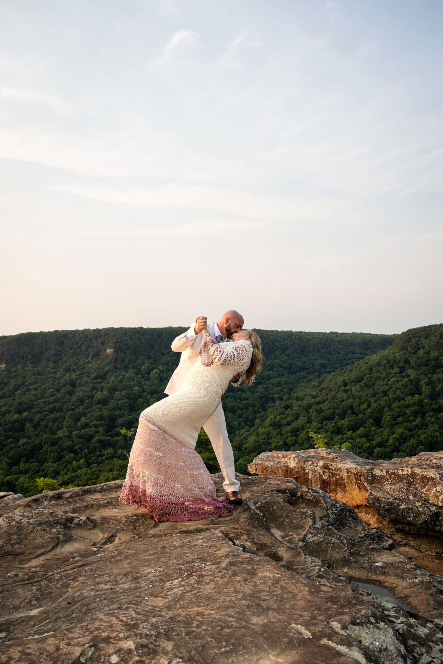 Couple embracing and kissing on a rocky mountain overlook with lush green valley views at sunset.