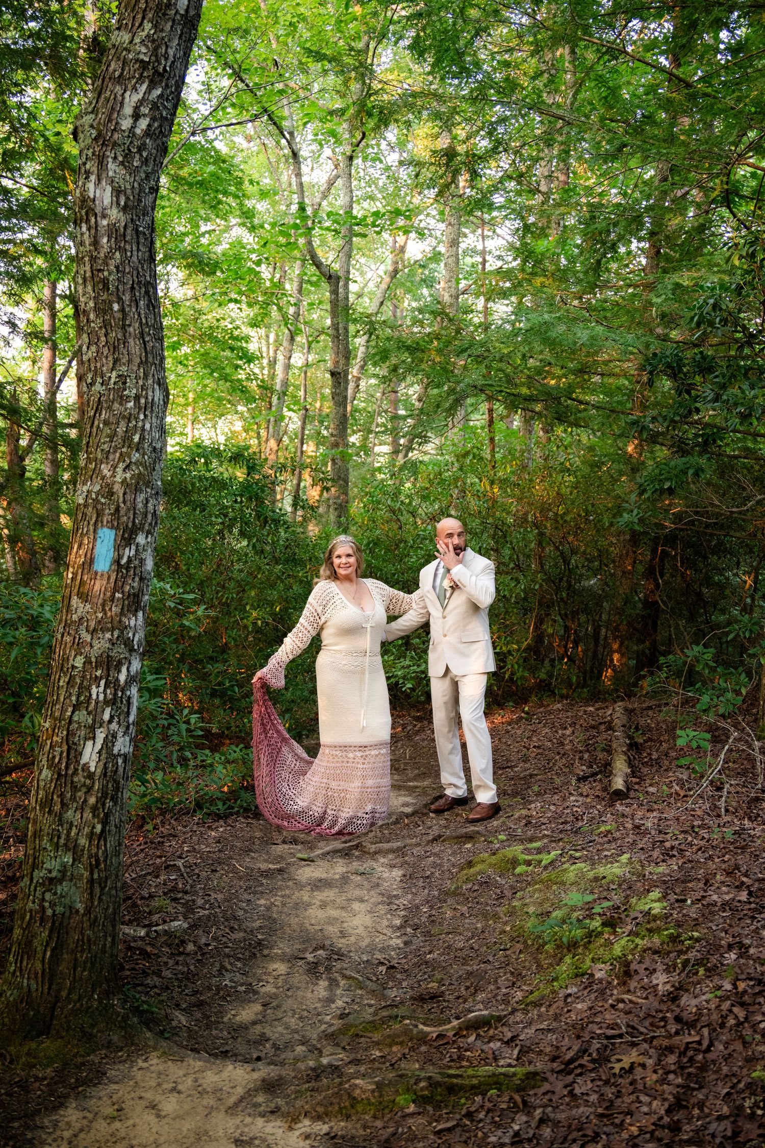 Couple in formal attire holding hands on a forest path, woman in pink dress, man in white suit, surrounded by lush green trees.