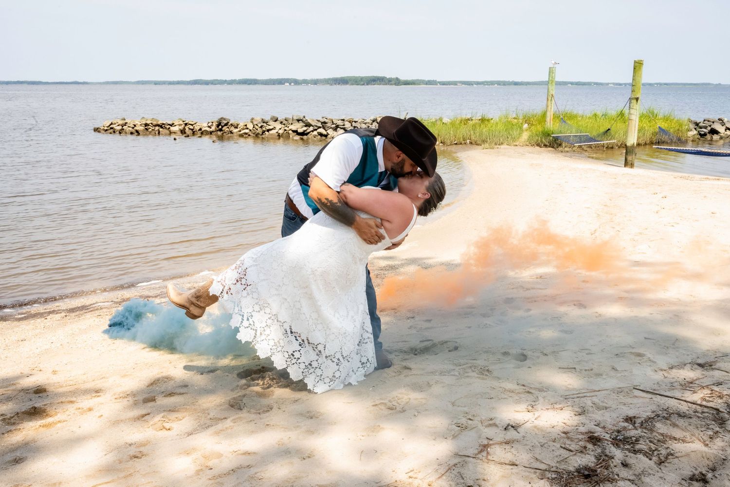 Bride and groom share a romantic dip kiss on sandy beach with colorful smoke bombs at waterfront wedding.