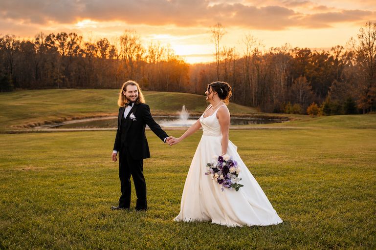 Bride and groom hold hands at sunset on a golf course, with a fountain pond and colorful sky in the background.