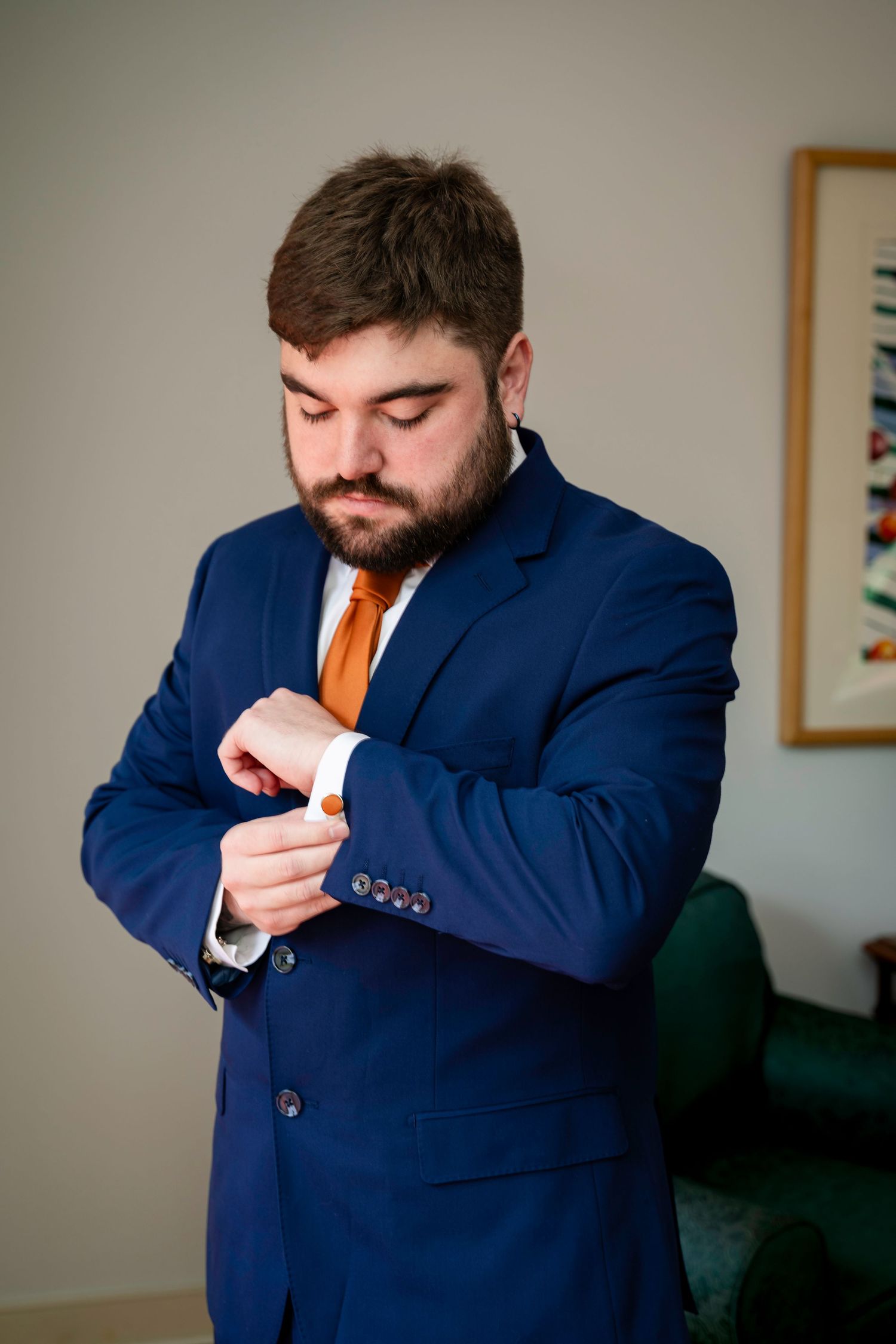 Dapper groom in navy blue suit and orange tie adjusting his cufflinks while getting ready for his wedding.