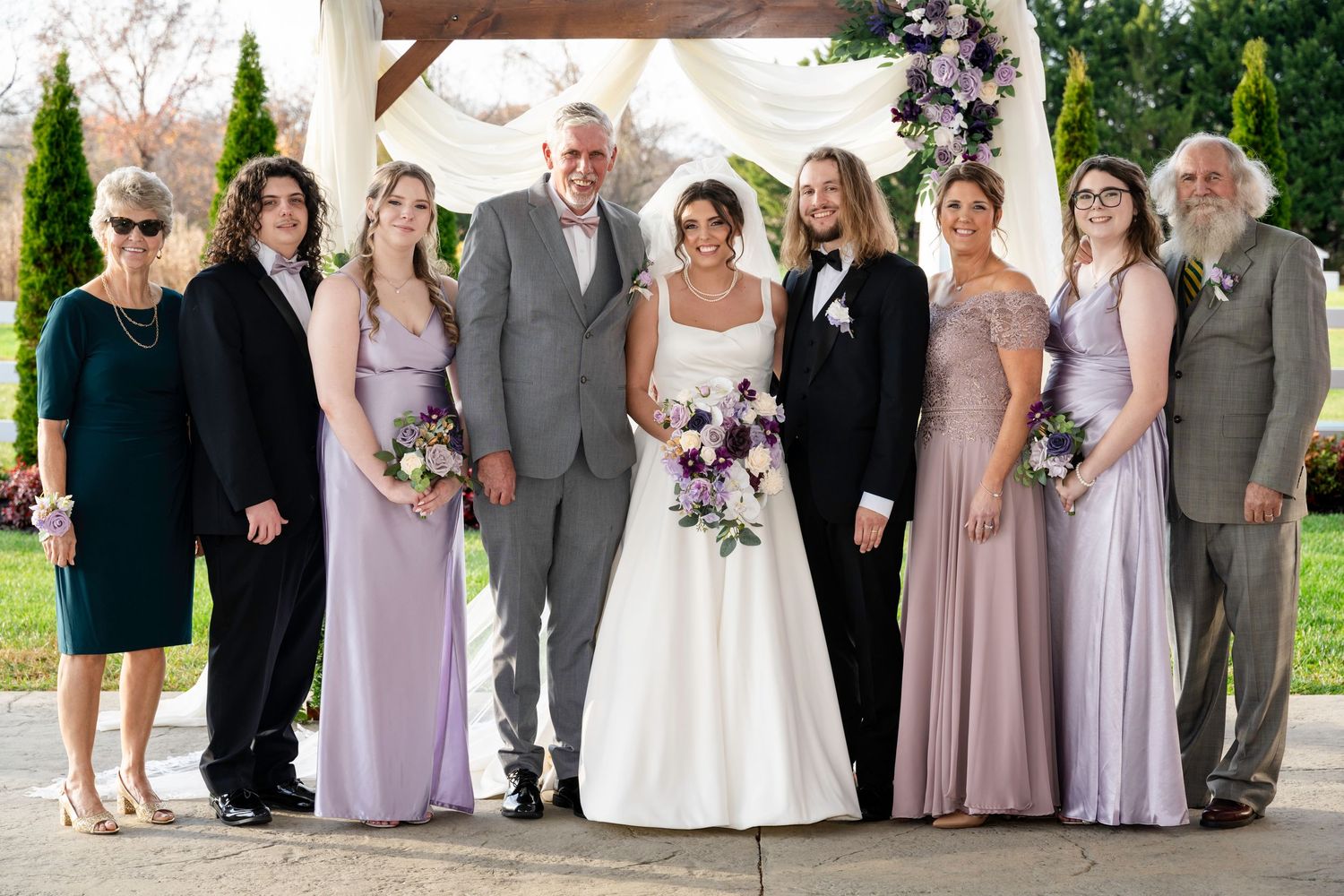 Bride and groom stand with family members in formal attire under a decorated outdoor pergola.