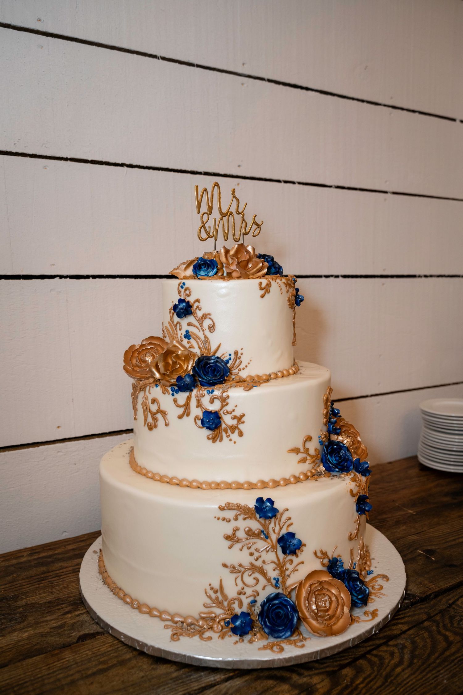 Three-tier white wedding cake decorated with gold lace piping and blue sugar roses topped with Mr and Mrs topper.