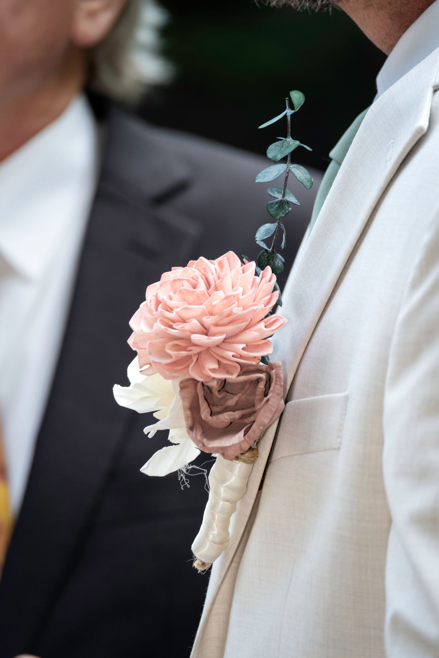 Groom in beige suit wearing a pink and mauve floral boutonniere with eucalyptus at a wedding.