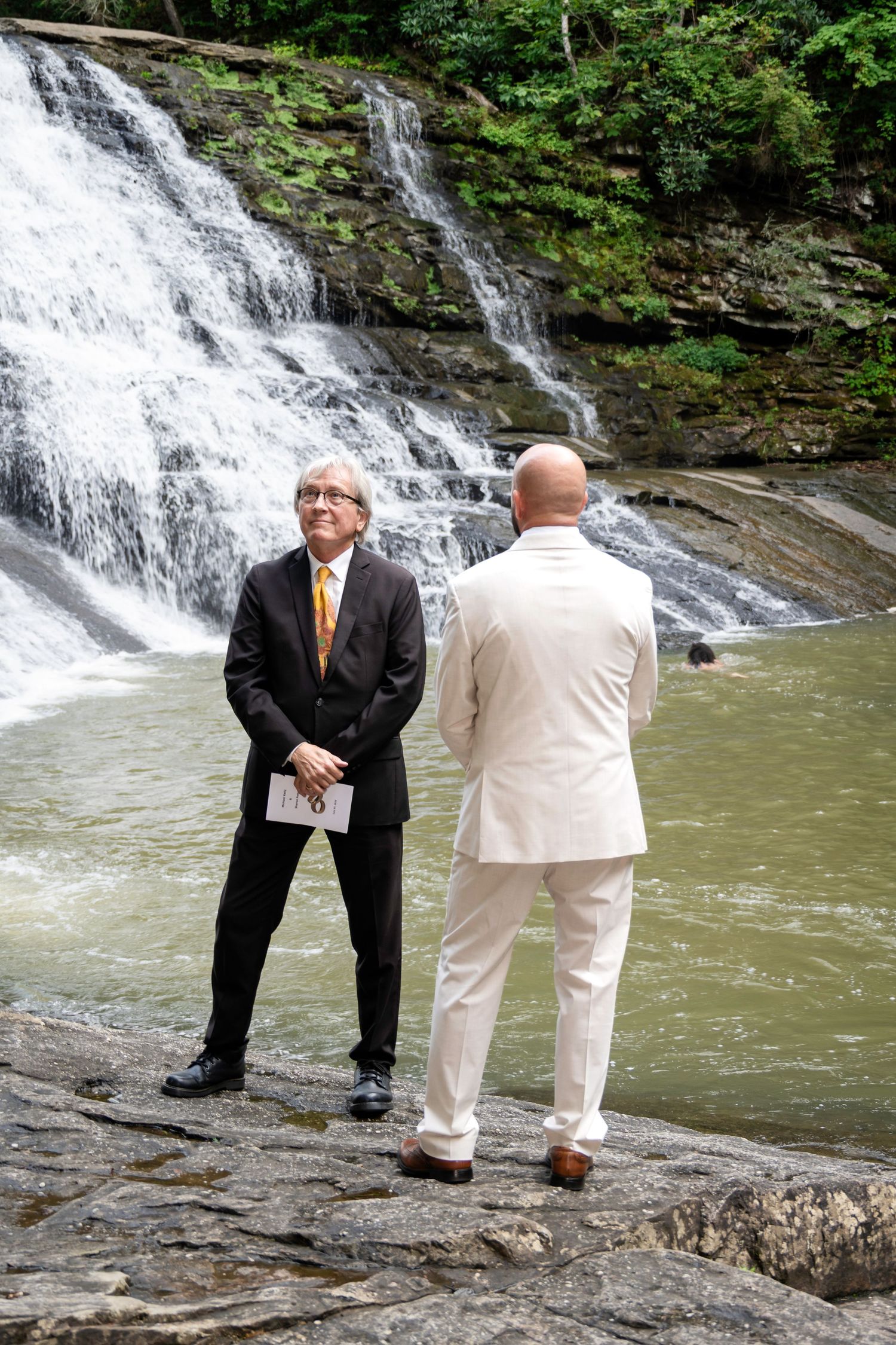 Two men in formal suits stand on rocks before a scenic waterfall during an outdoor wedding ceremony.