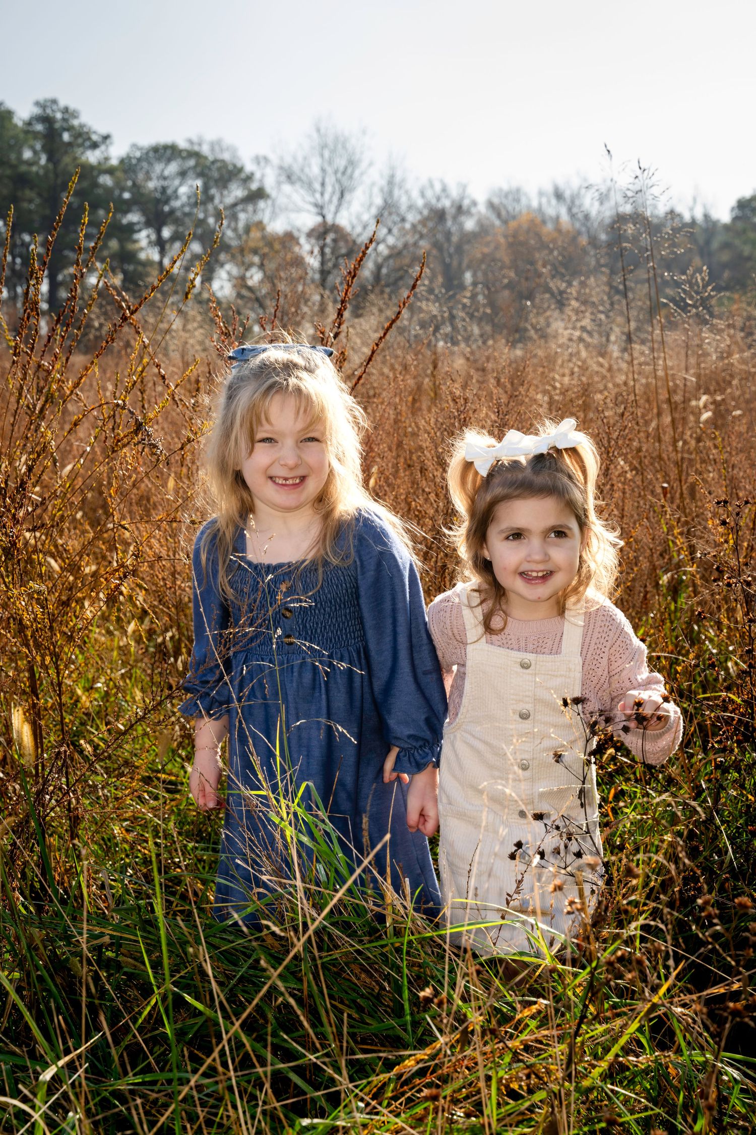 Two young girls smiling in a sunlit autumn field with tall golden grasses and trees in the background.  Fall Family Session