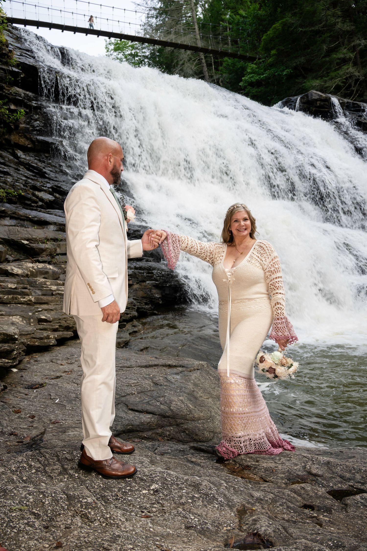 Couple in wedding attire holding hands before a cascading waterfall, bride in lace gown with bouquet, groom in cream suit.