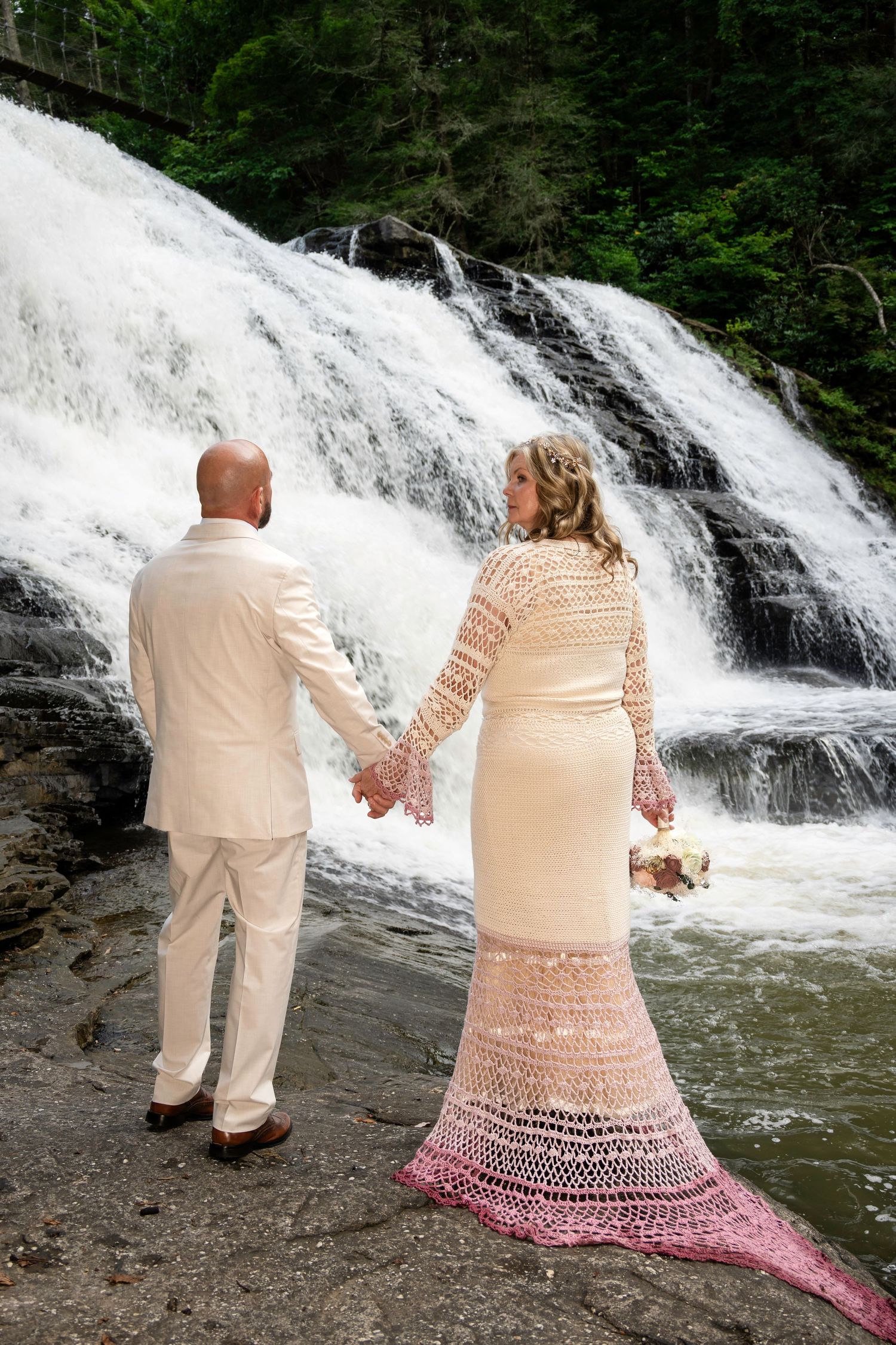 Bride in pink crochet gown and groom in white suit hold hands before a stunning waterfall backdrop.