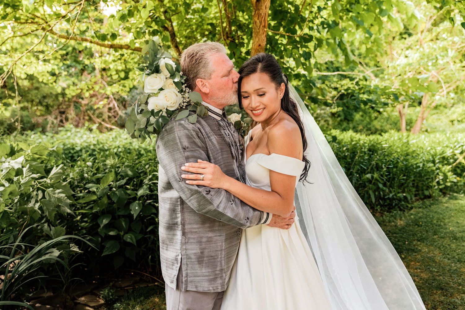 Bride and groom embrace outdoors in lush garden, bride in off-shoulder gown with veil, groom in gray suit.