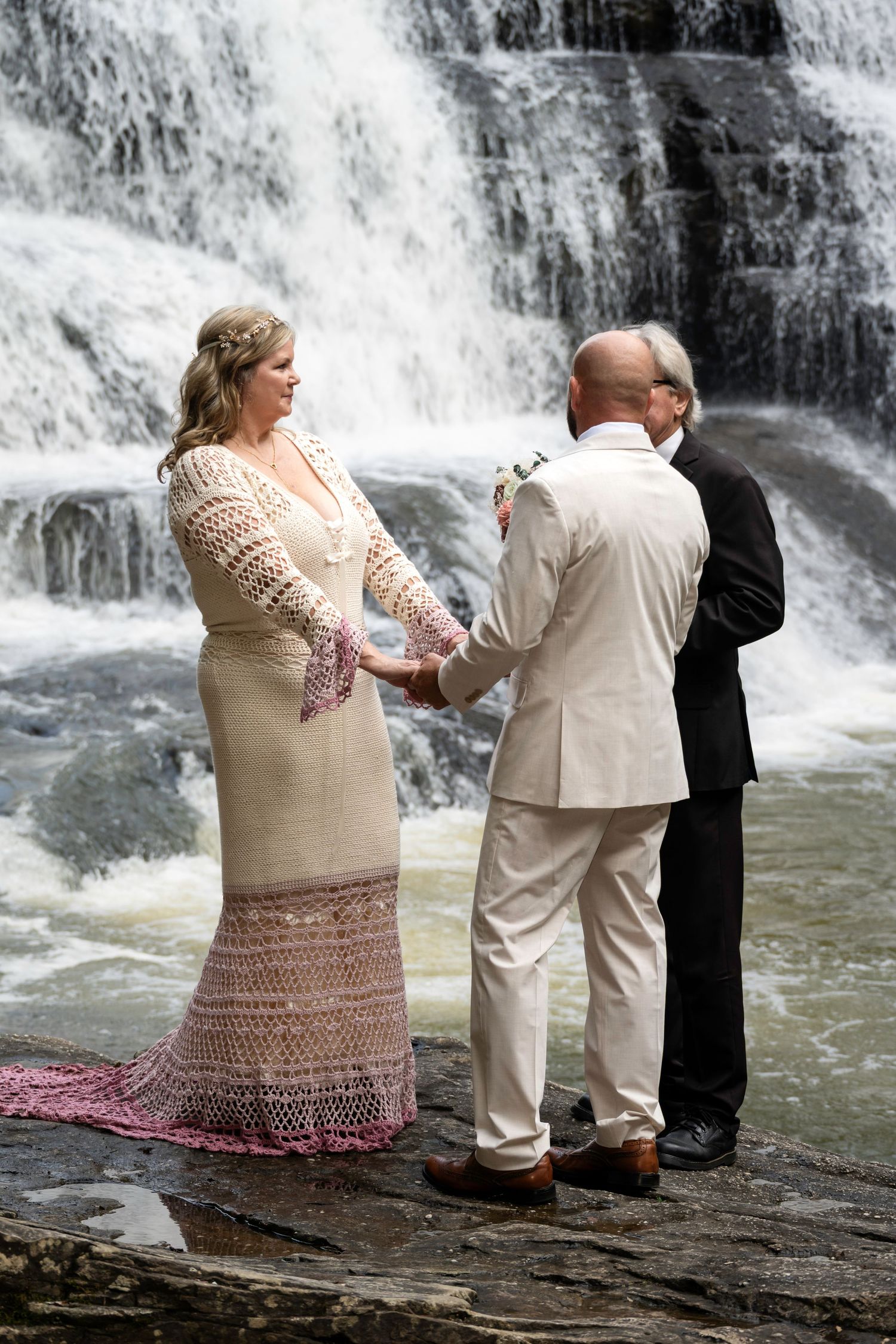 Couple exchanges vows in front of stunning waterfall during outdoor wedding ceremony, bride in lace gown, groom in cream suit.