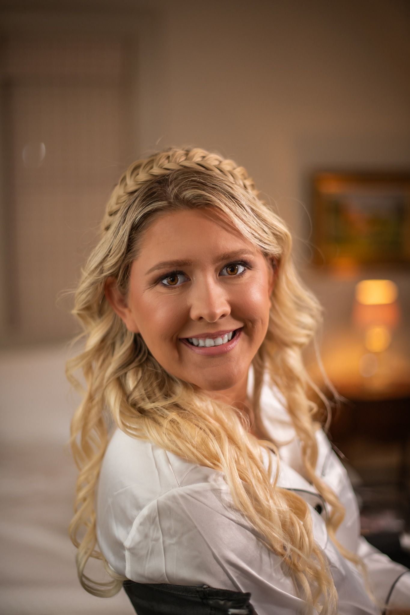 Smiling blonde bride with braided crown hairstyle wearing white robe in warmly lit room. Maryland Wedding Photography