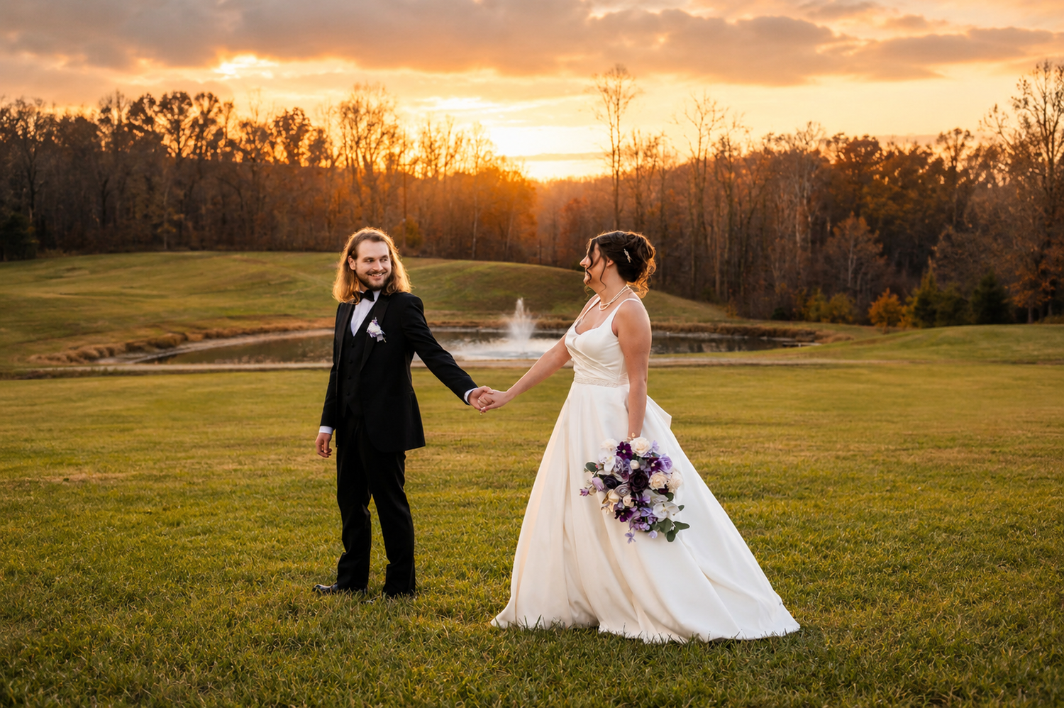 Bride and groom hold hands at sunset on a lush green lawn with a pond and fountain backdrop.