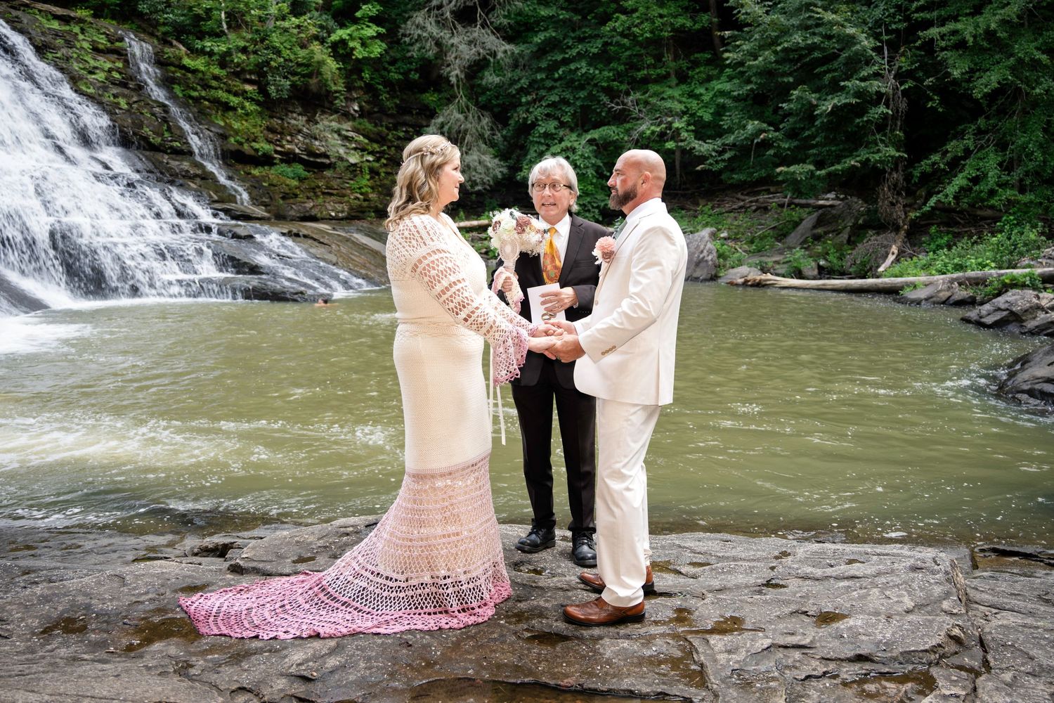 Couple exchanges vows before a waterfall with an officiant, bride in pink lace gown, groom in white suit.