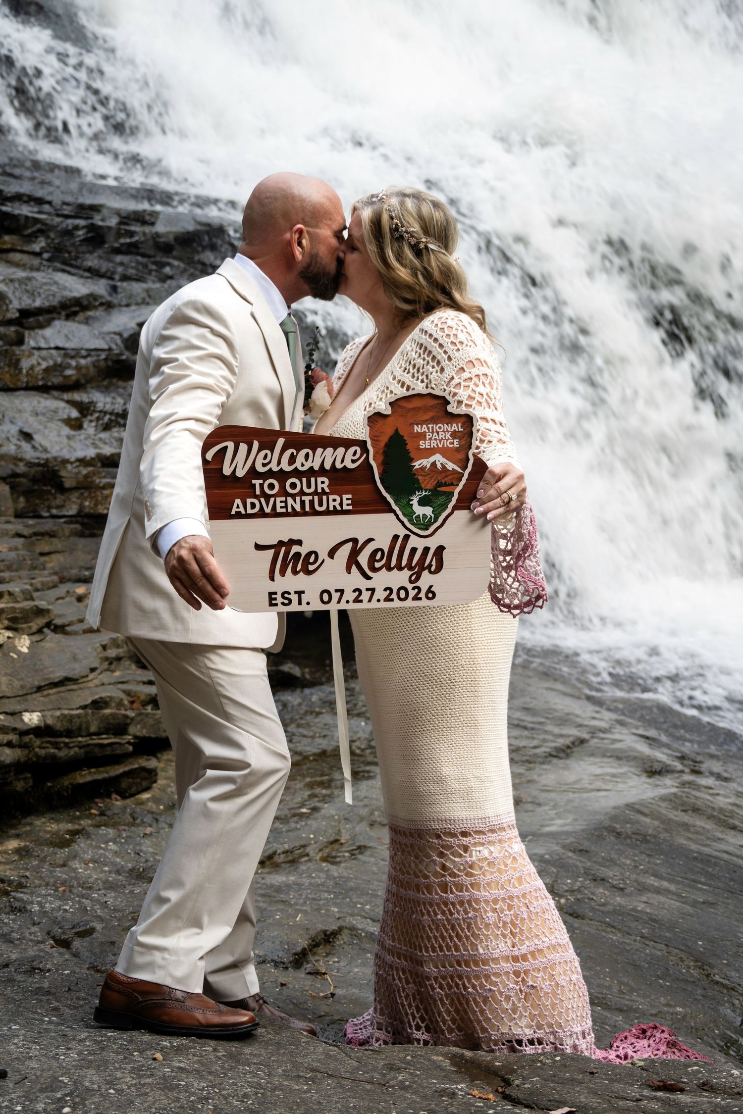 Newlyweds the Kellys kiss before a waterfall holding a welcome sign, bride in crochet gown, groom in white suit.