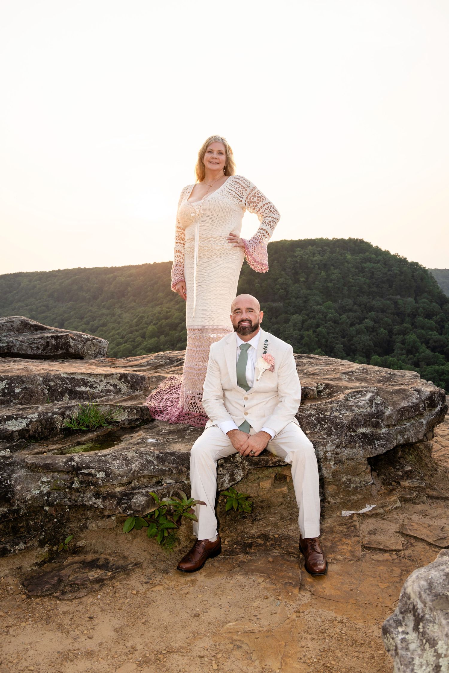 Couple in wedding attire poses on rocky overlook with lush green mountains at sunset.
