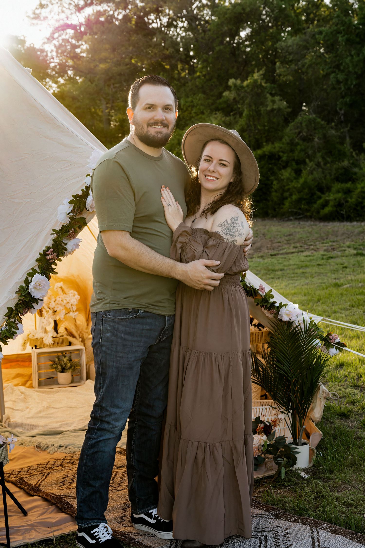 A couple embraces outdoors at golden hour near a boho tent with floral decor, woman wearing a hat and maxi dress.