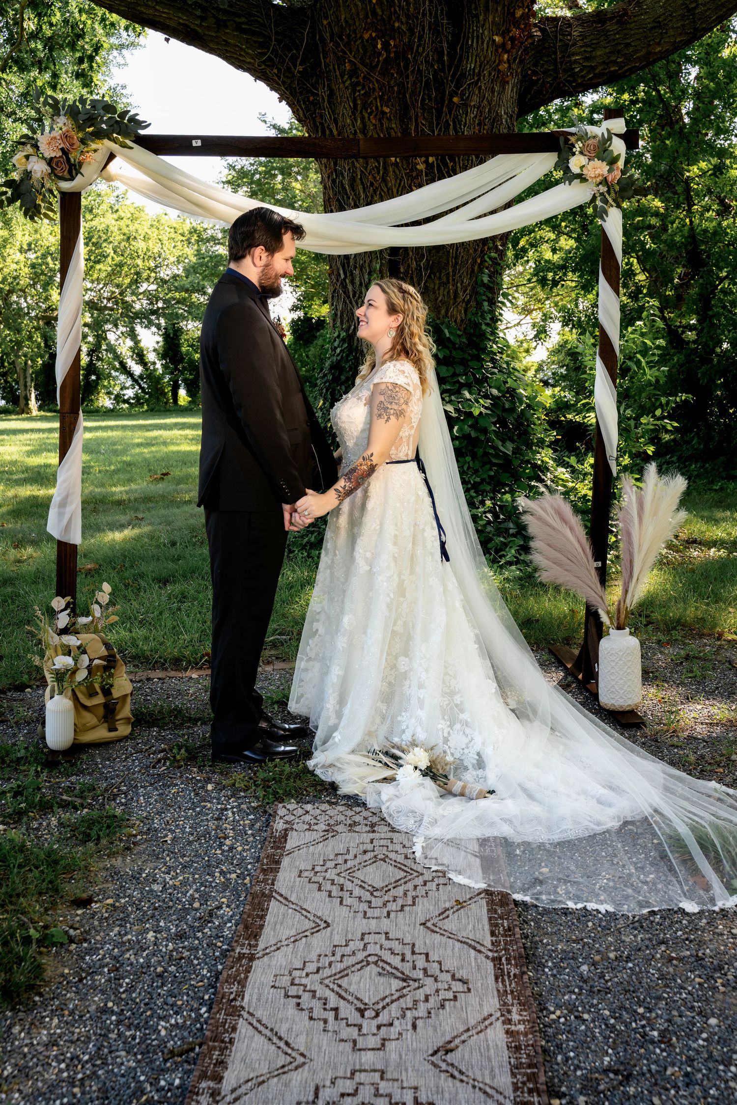 Bride and groom holding hands under a decorated outdoor wedding arch surrounded by lush green trees.