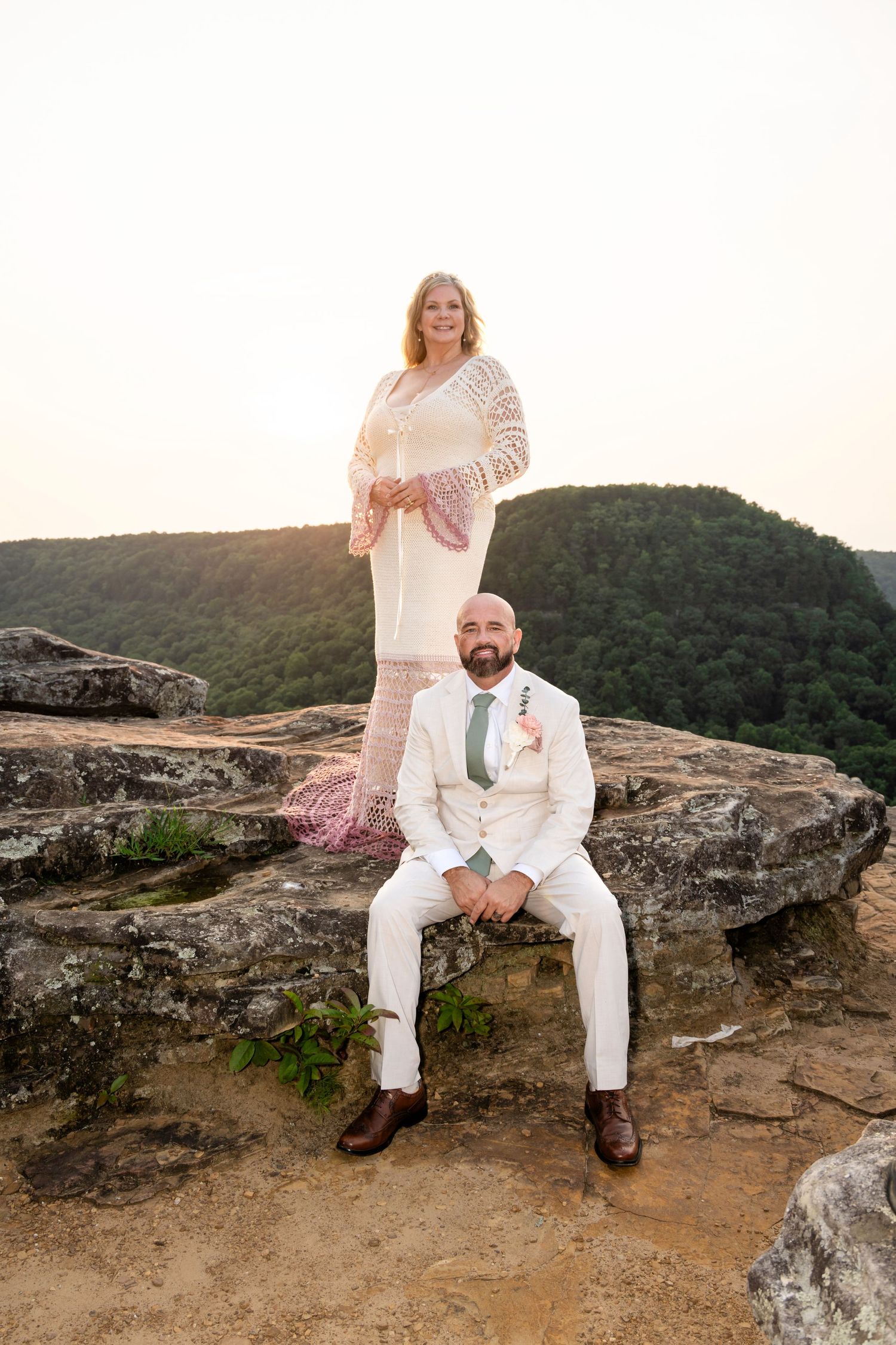 Bride and groom posing on rocky mountain overlook at sunset, groom seated in cream suit, bride standing in white dress.