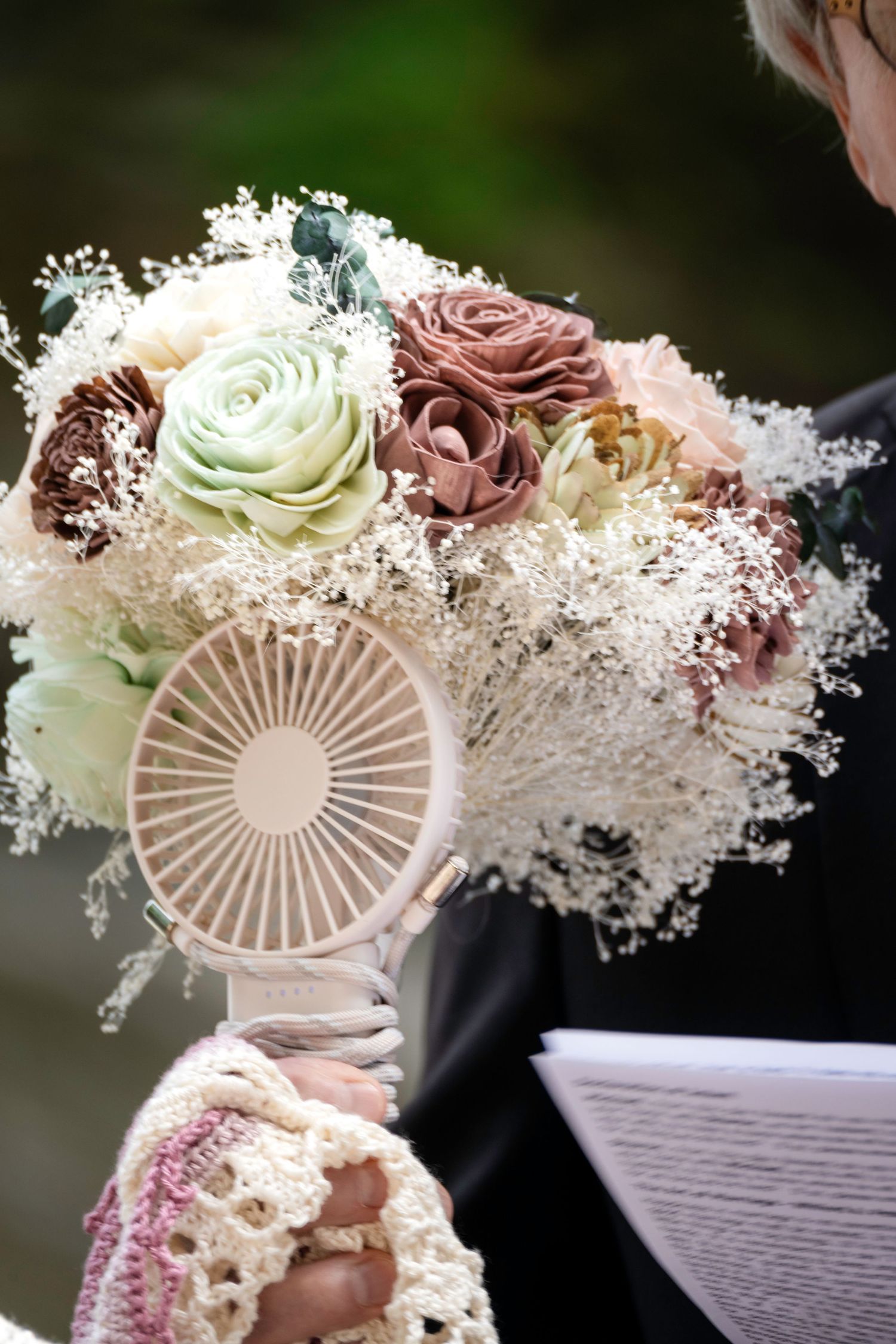 Bride holding a pastel wedding bouquet with roses and baby's breath alongside a small handheld fan outdoors.