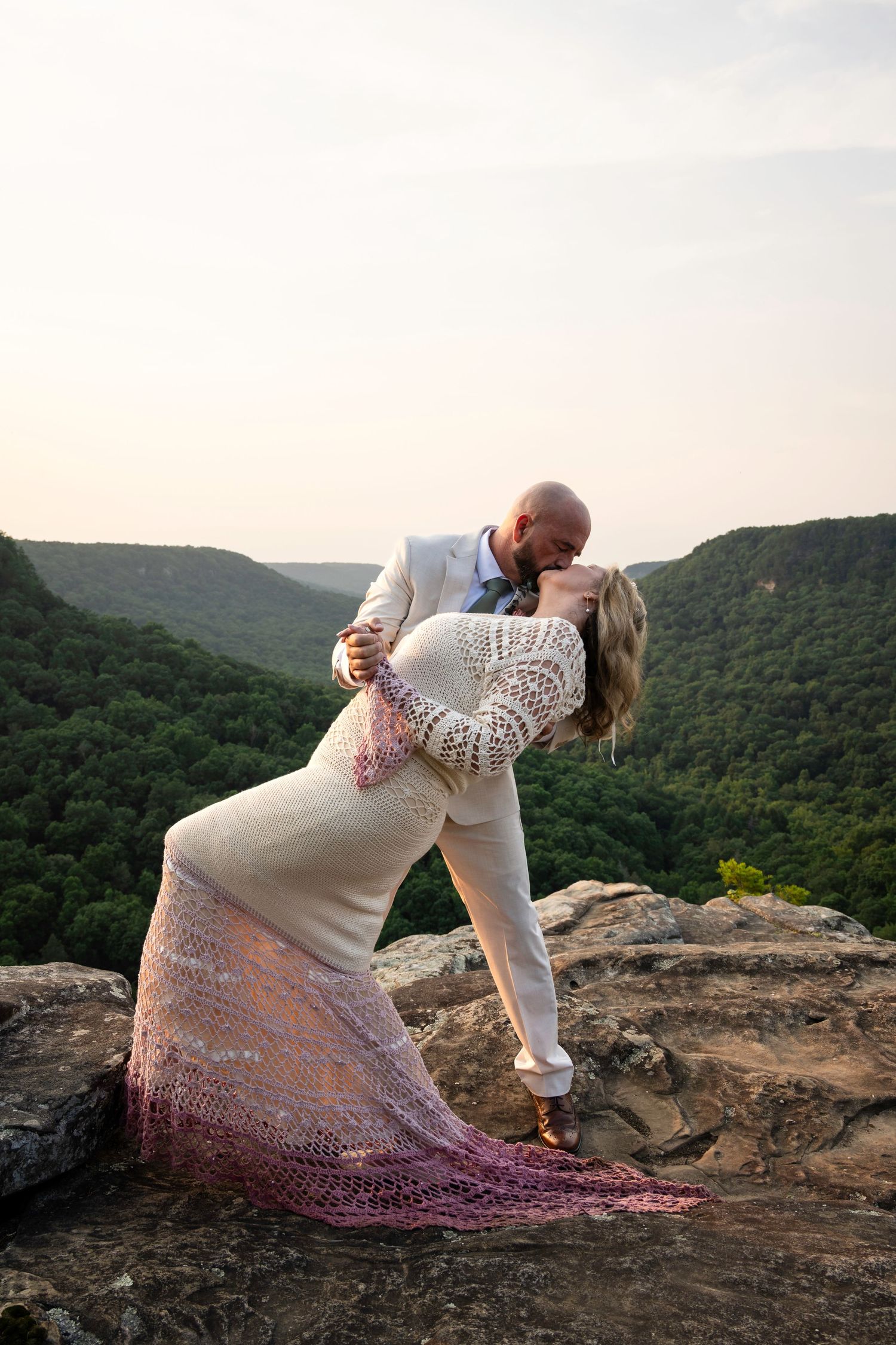 Bride and groom share a romantic dip kiss on a rocky mountain overlook surrounded by lush green valleys at sunset.