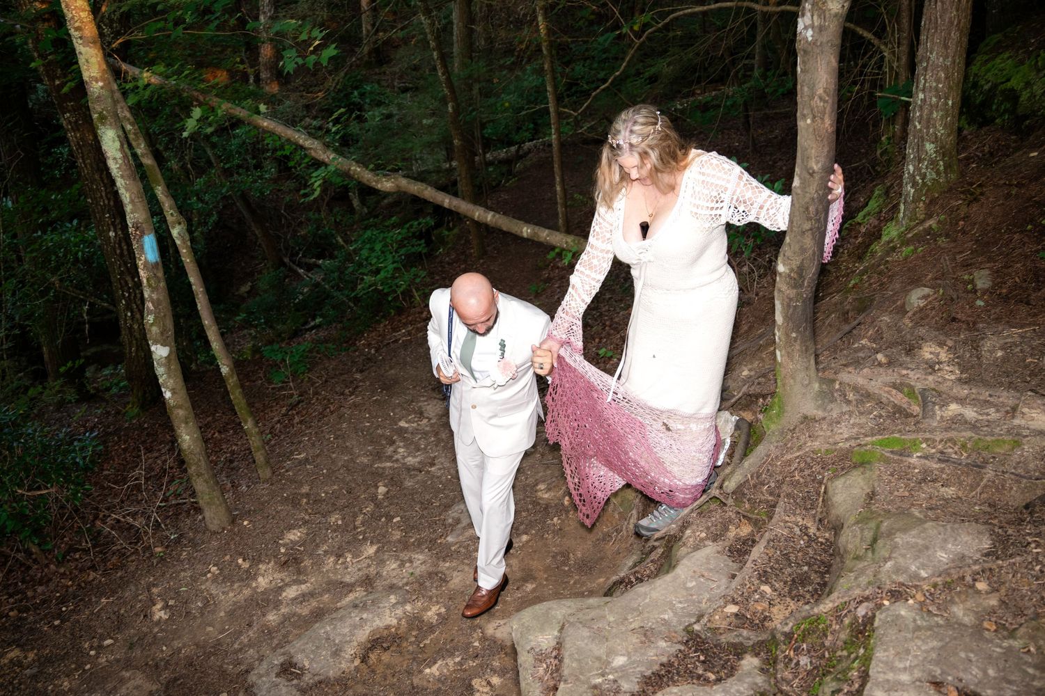 Bride and groom in white attire climbing rocky forest trail, woman holding pink dress while man assists her up stone steps.
