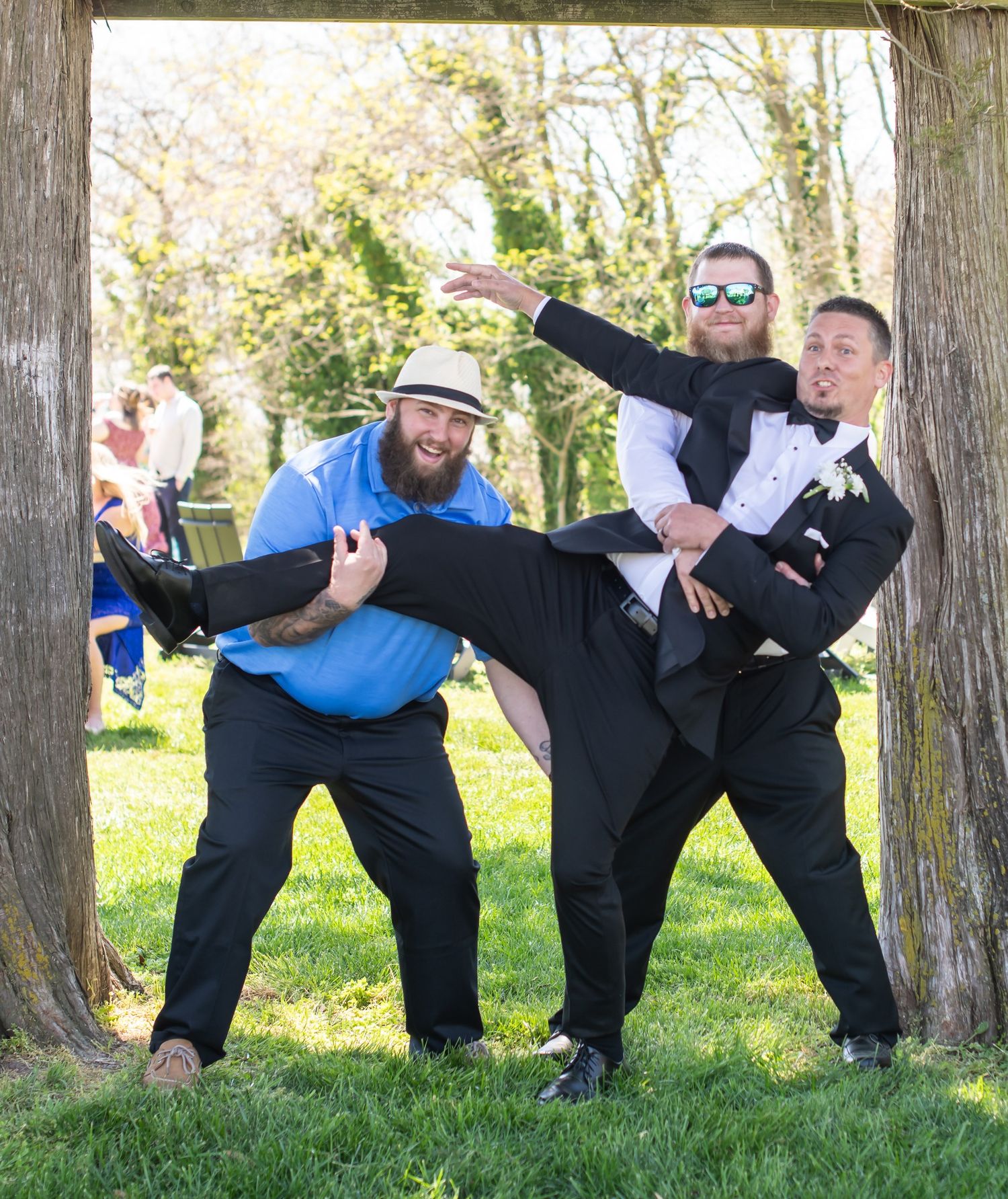 Three groomsmen strike a fun playful pose under a rustic wooden arch at an outdoor wedding.