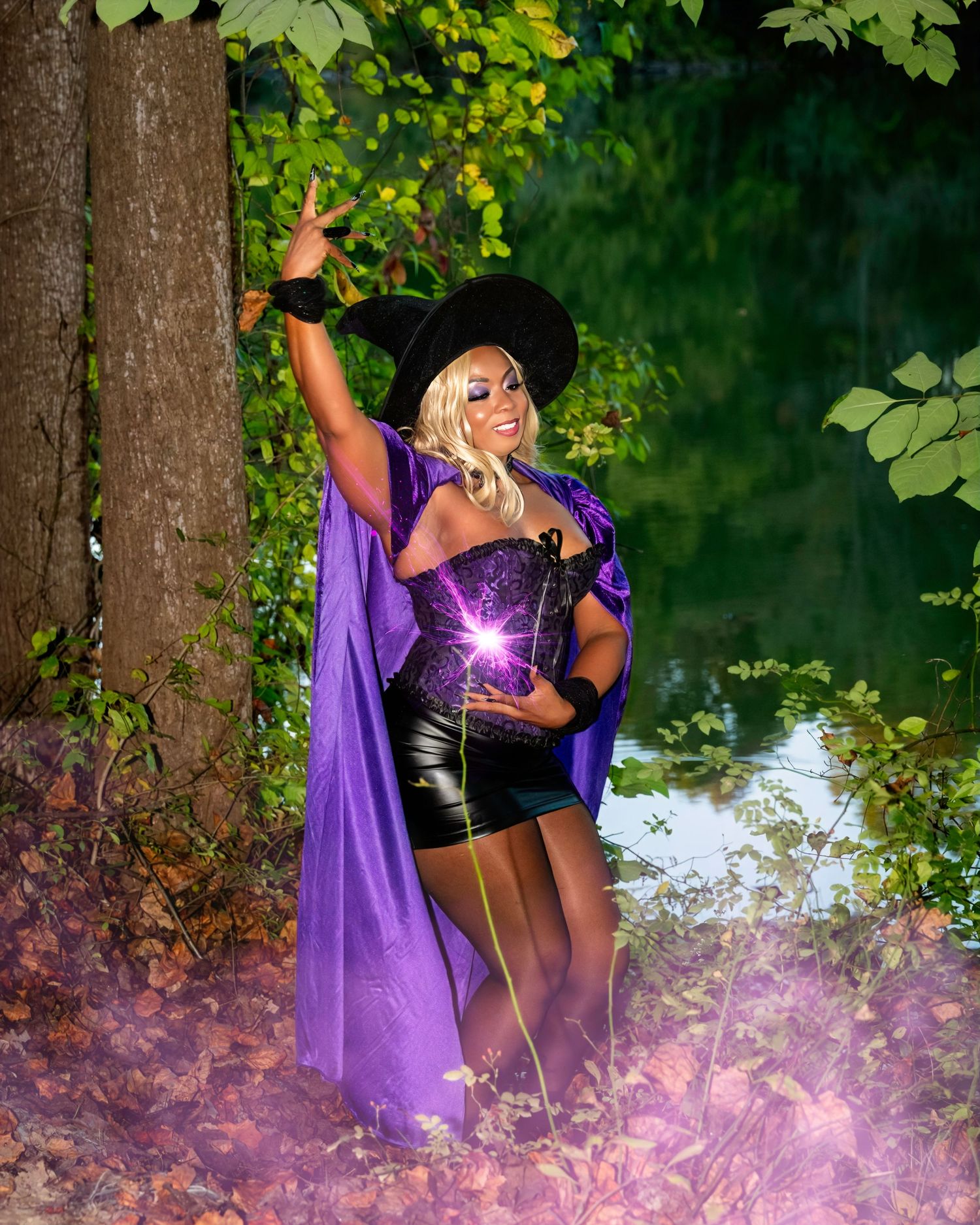 A woman in a purple witch costume with cape and hat poses dramatically in a forest with magical glowing effects.