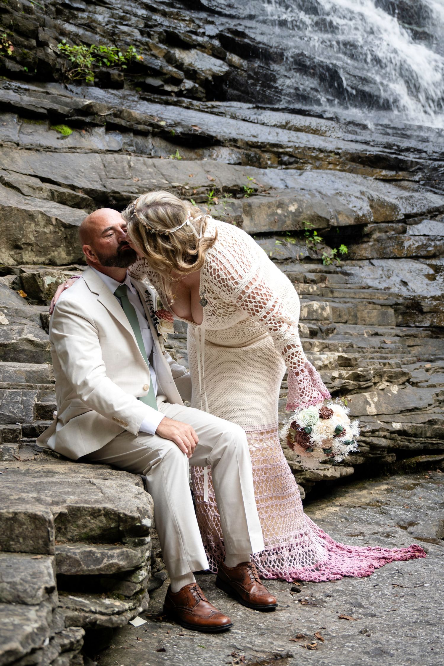 Bride in crochet dress kisses groom in white suit on stone steps before a waterfall.