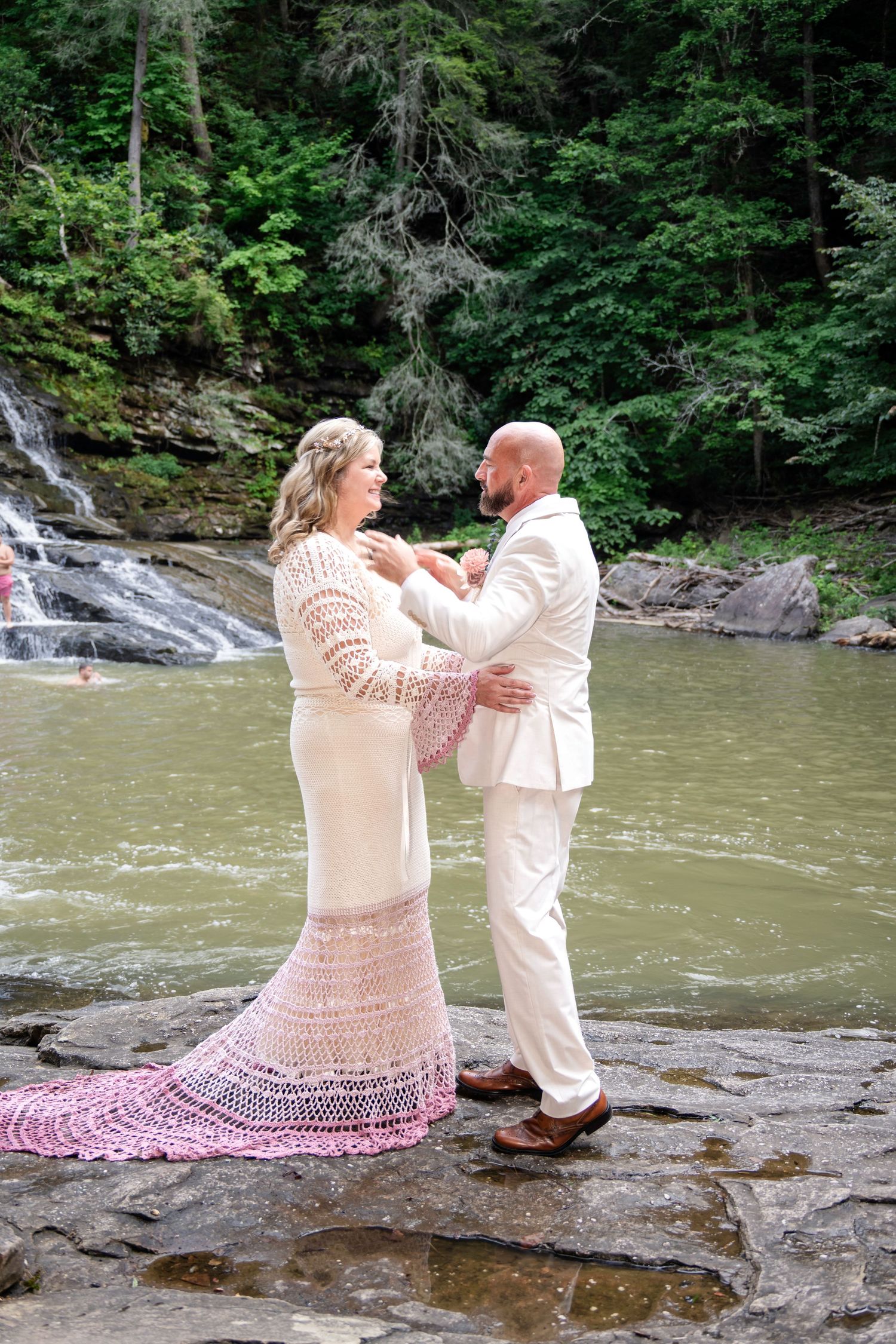 Bride in crochet dress and groom in white suit dance on rocks by a waterfall in a lush forest wedding.