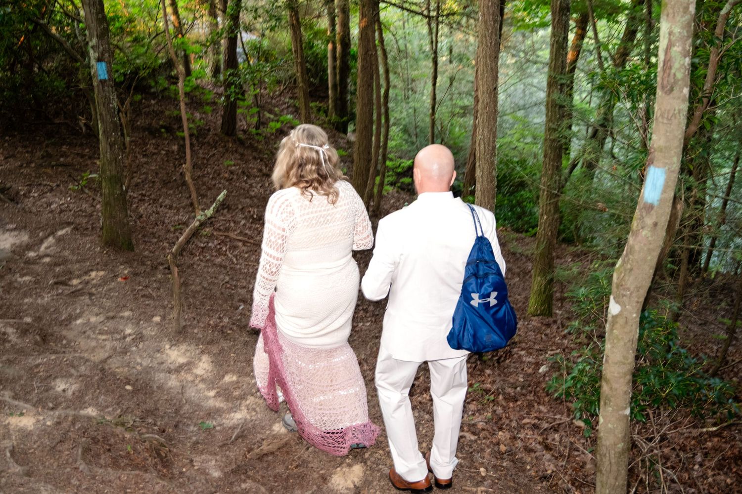 Couple in white wedding attire walking hand-in-hand along a forest trail surrounded by tall trees.