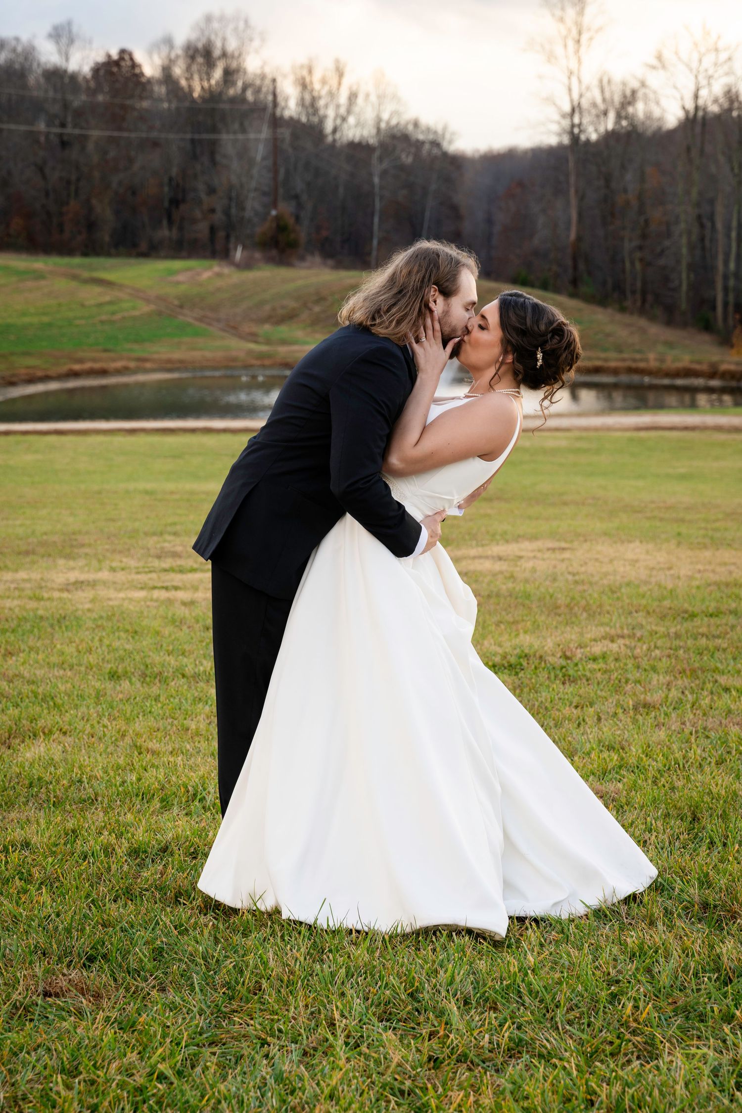 Groom dips bride for romantic kiss on green lawn with pond in background at sunset.