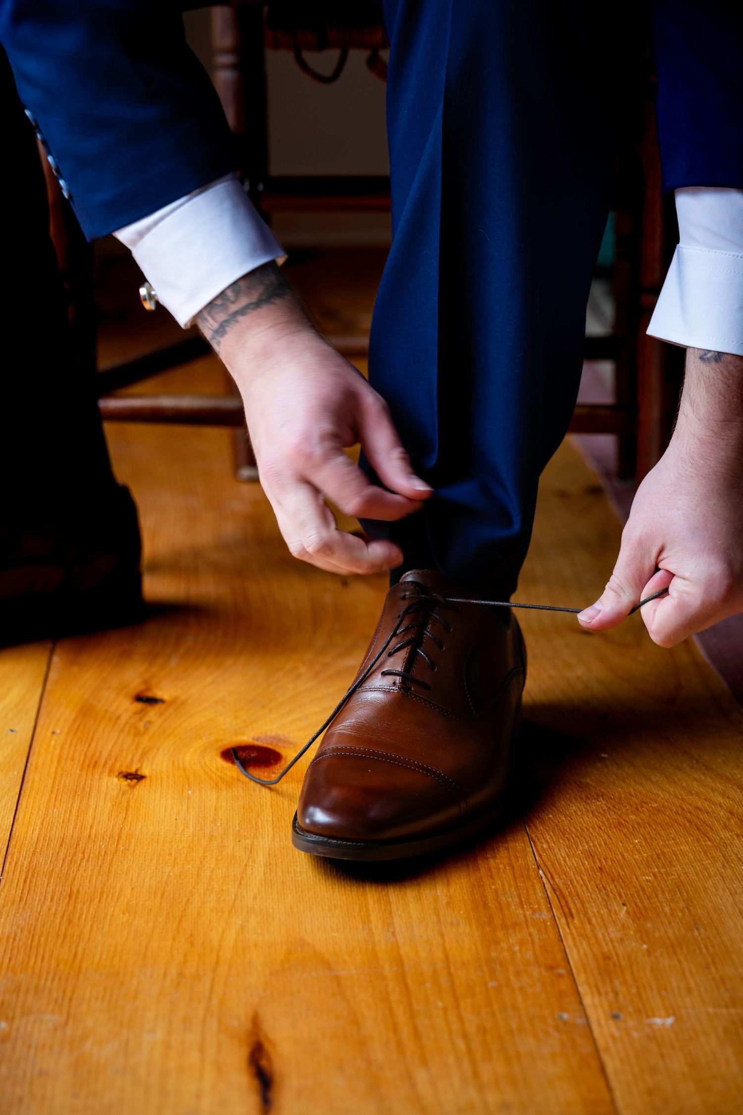 Groom in navy blue suit tying brown leather dress shoes on a warm hardwood floor before the wedding ceremony.