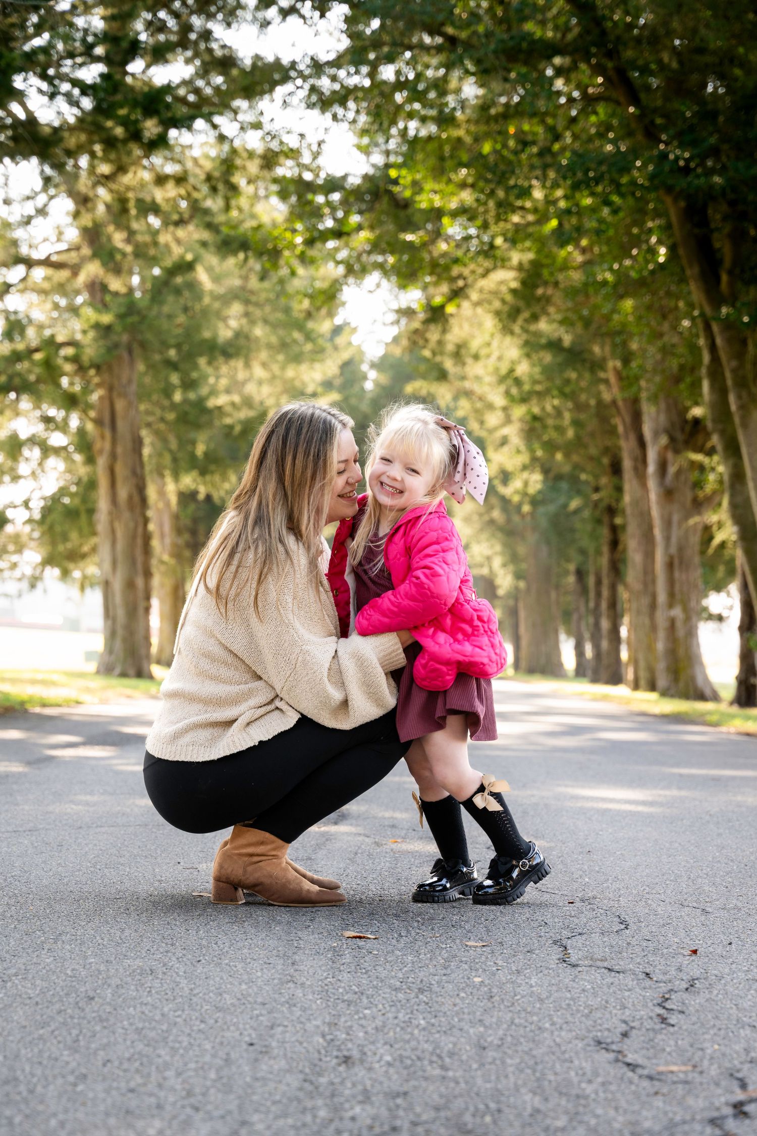 A mother crouches down to hug her laughing toddler daughter in a pink coat on a tree-lined path.
