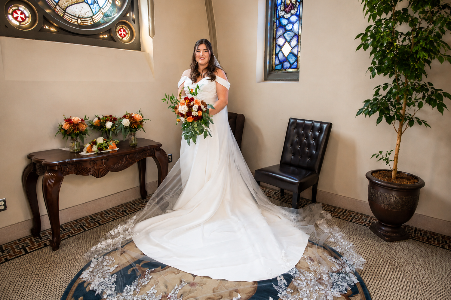 Bride in white gown with lace train holds colorful bouquet beside stained glass windows in elegant venue.