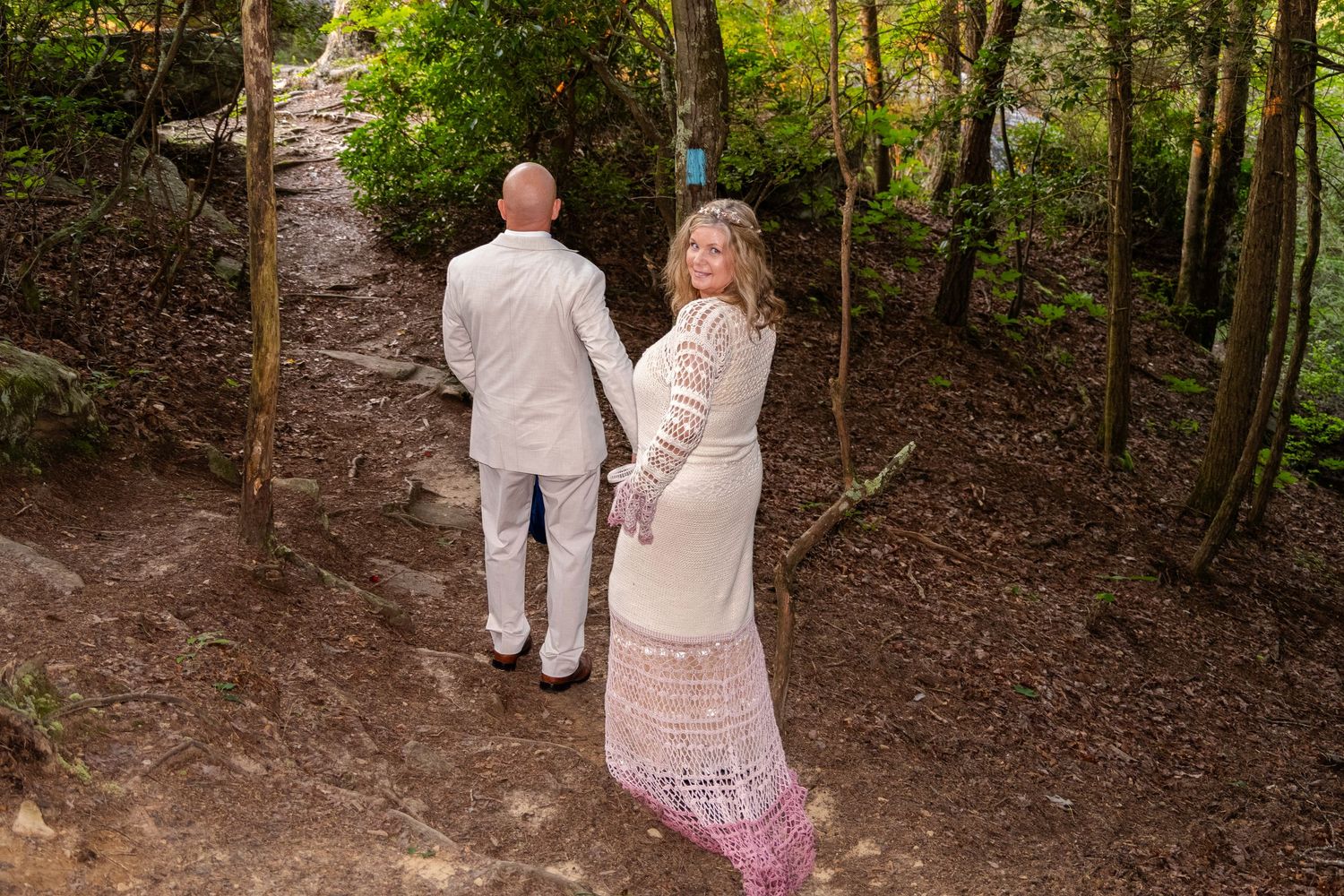 Couple in bohemian wedding attire holding hands on a forest trail, bride in lace dress looking back over shoulder.