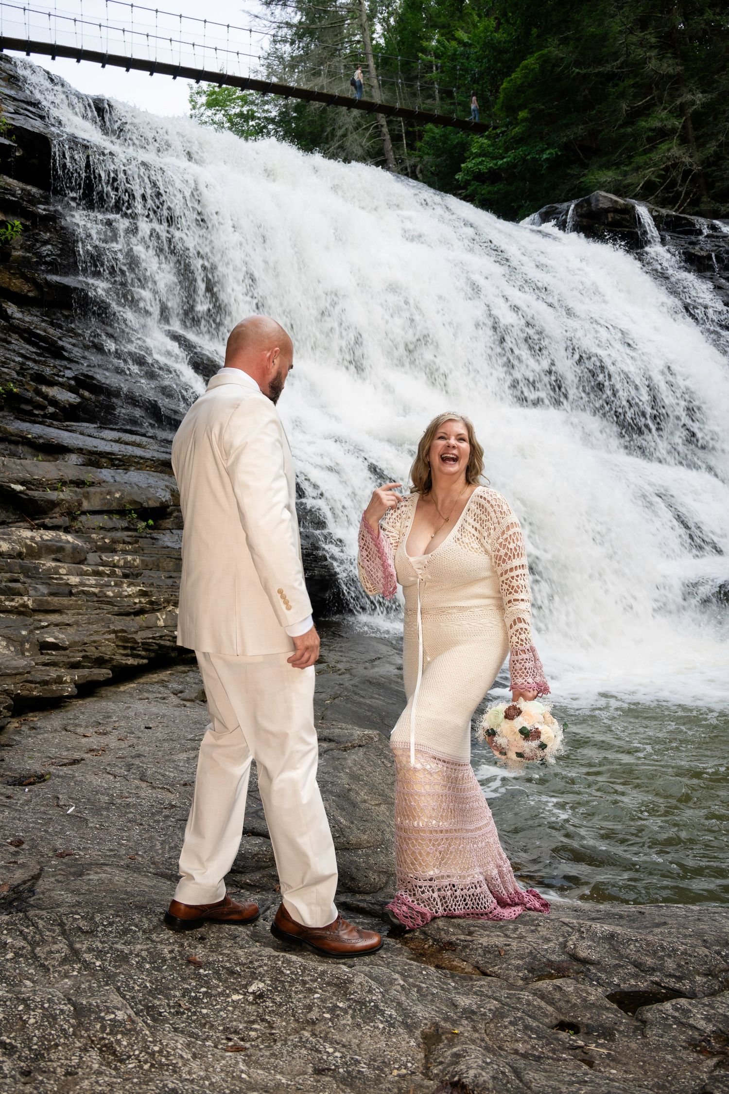 Laughing bride in white crochet gown holds bouquet with groom in cream suit before stunning waterfall backdrop.