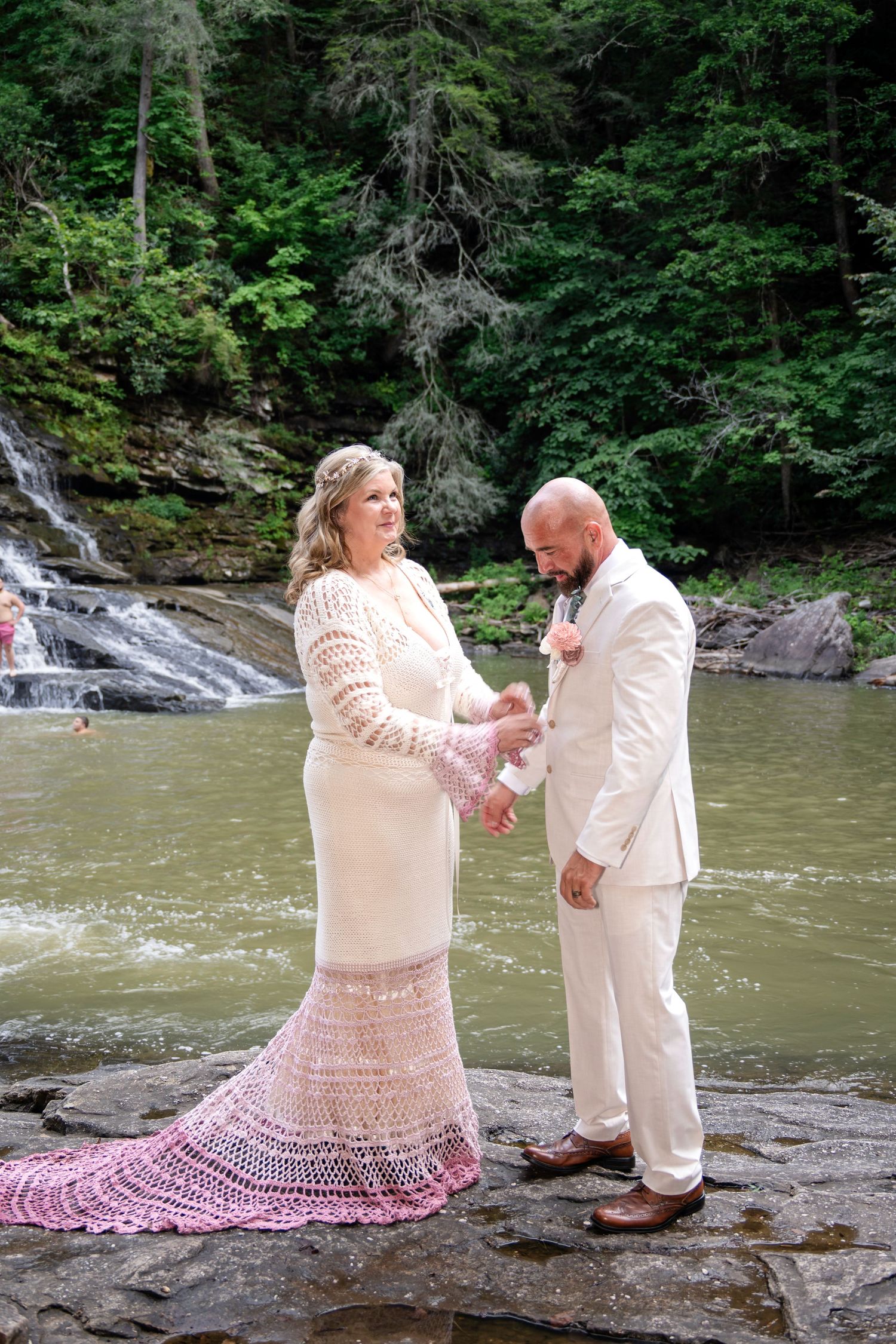 Bride in white crochet gown and groom in cream suit exchange rings on rocks by a waterfall.