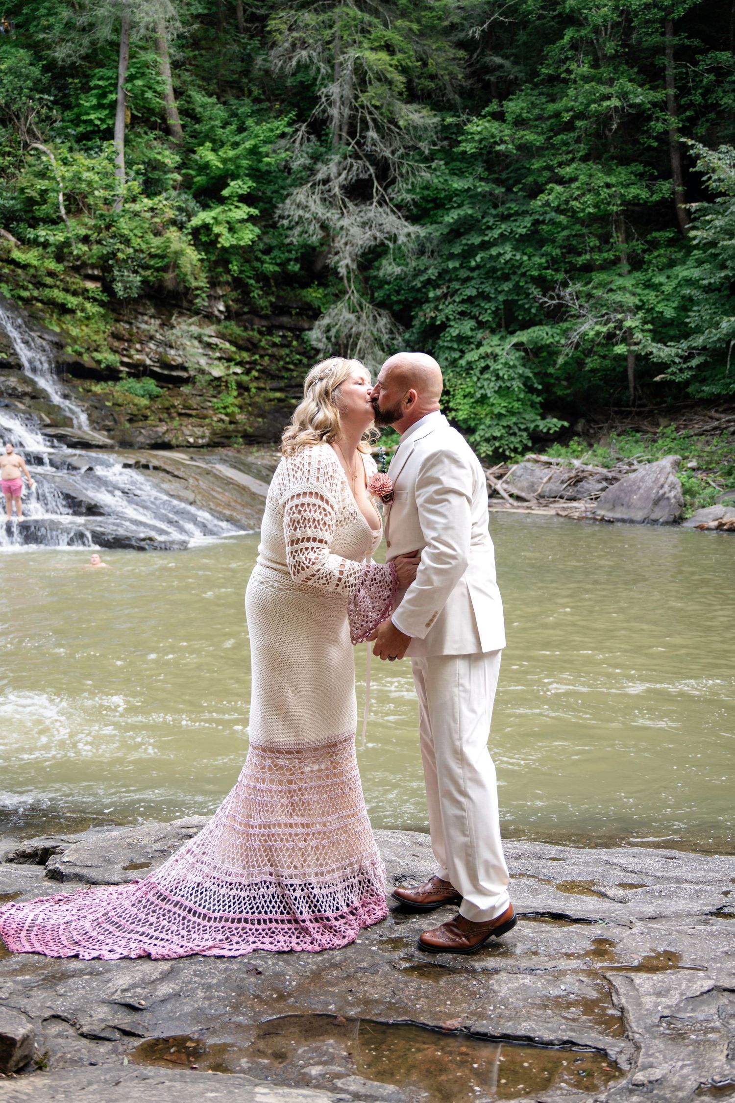 Couple sharing a romantic moment in wedding attire by a waterfall, bride in pink crochet gown, groom in tan suit.