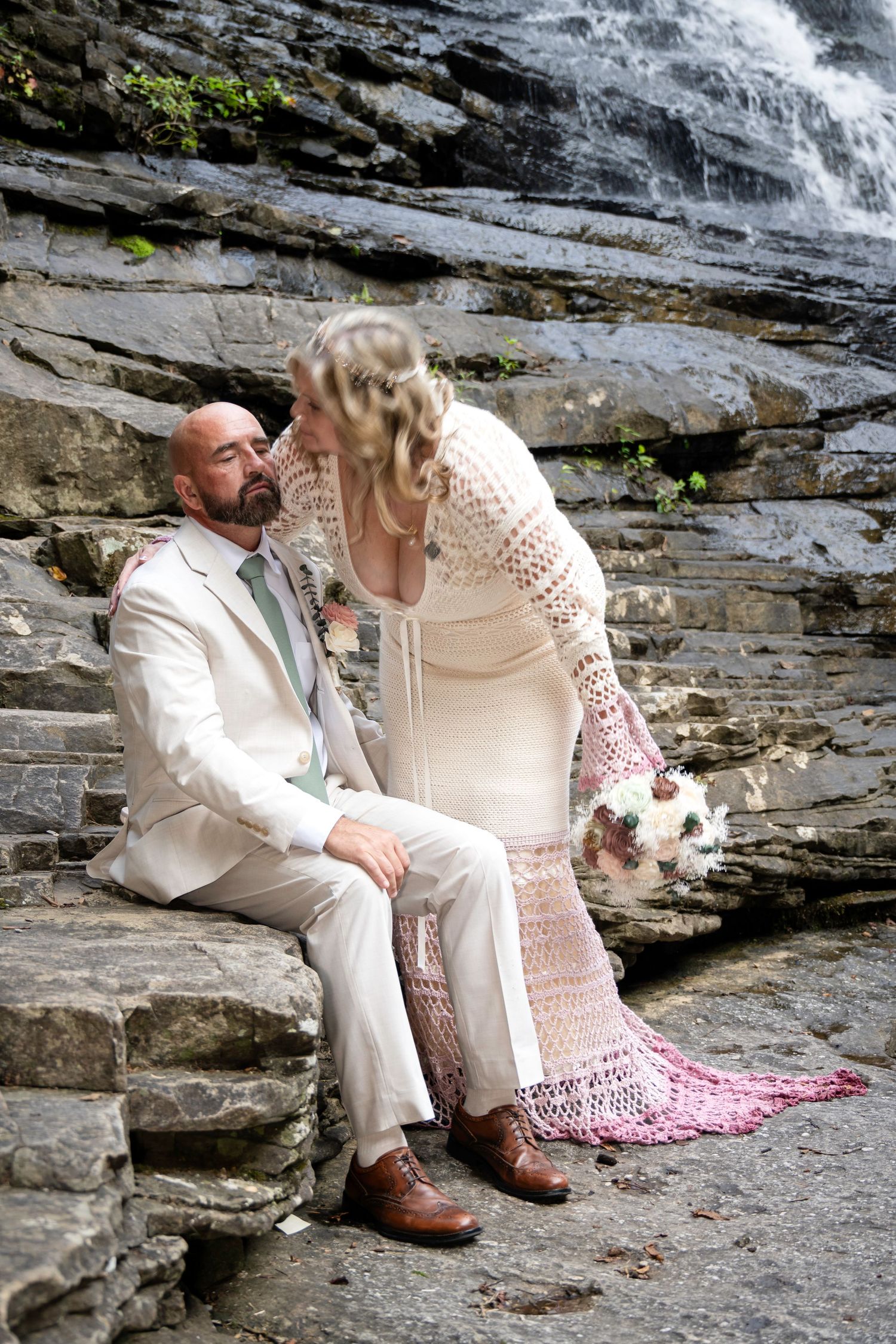 Bride kissing bald groom on stone steps near waterfall, wedding attire with crochet dress and bouquet.
