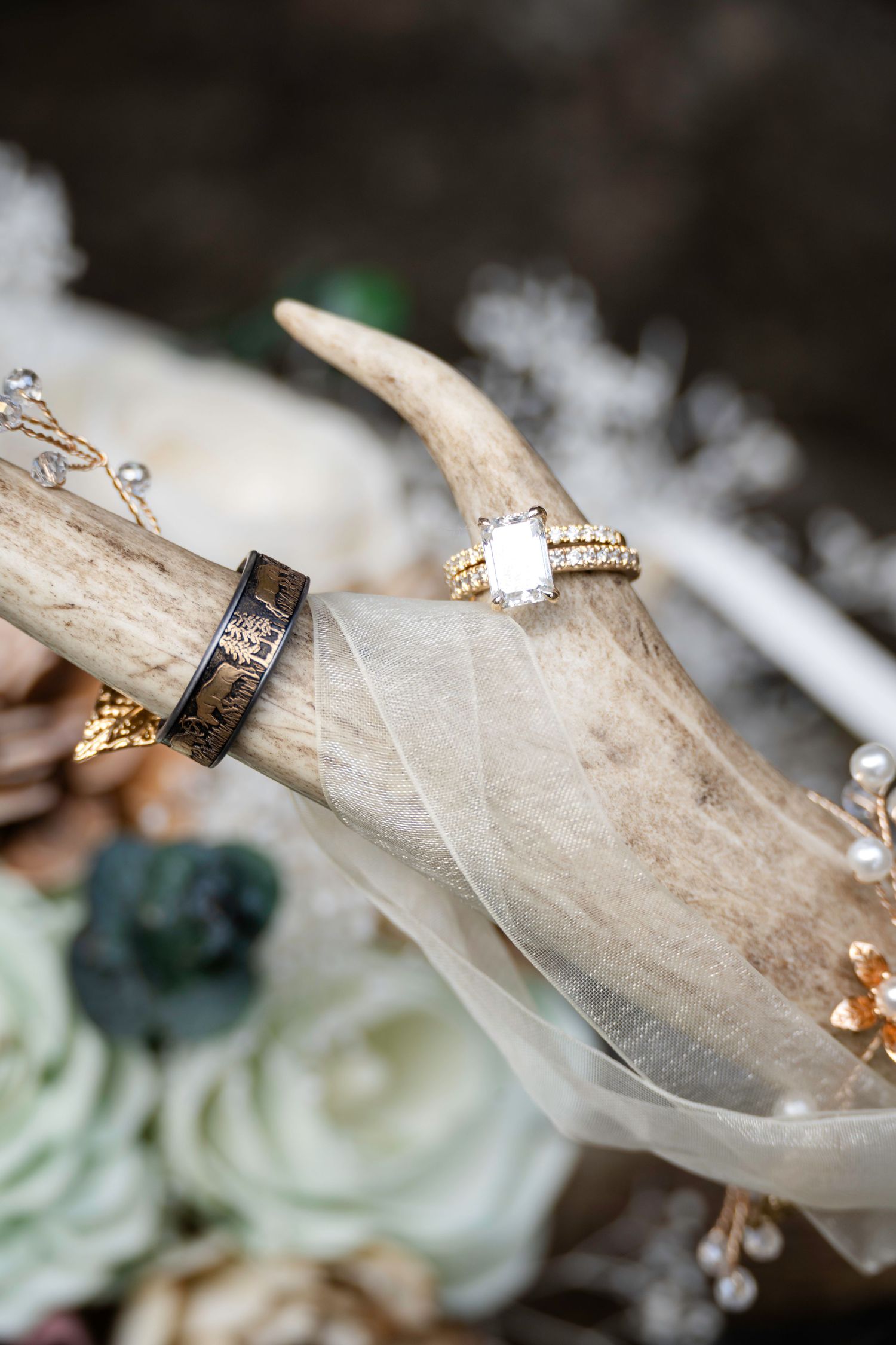 Gold diamond engagement ring and black band displayed on antler with white roses and tulle in wedding flat lay.