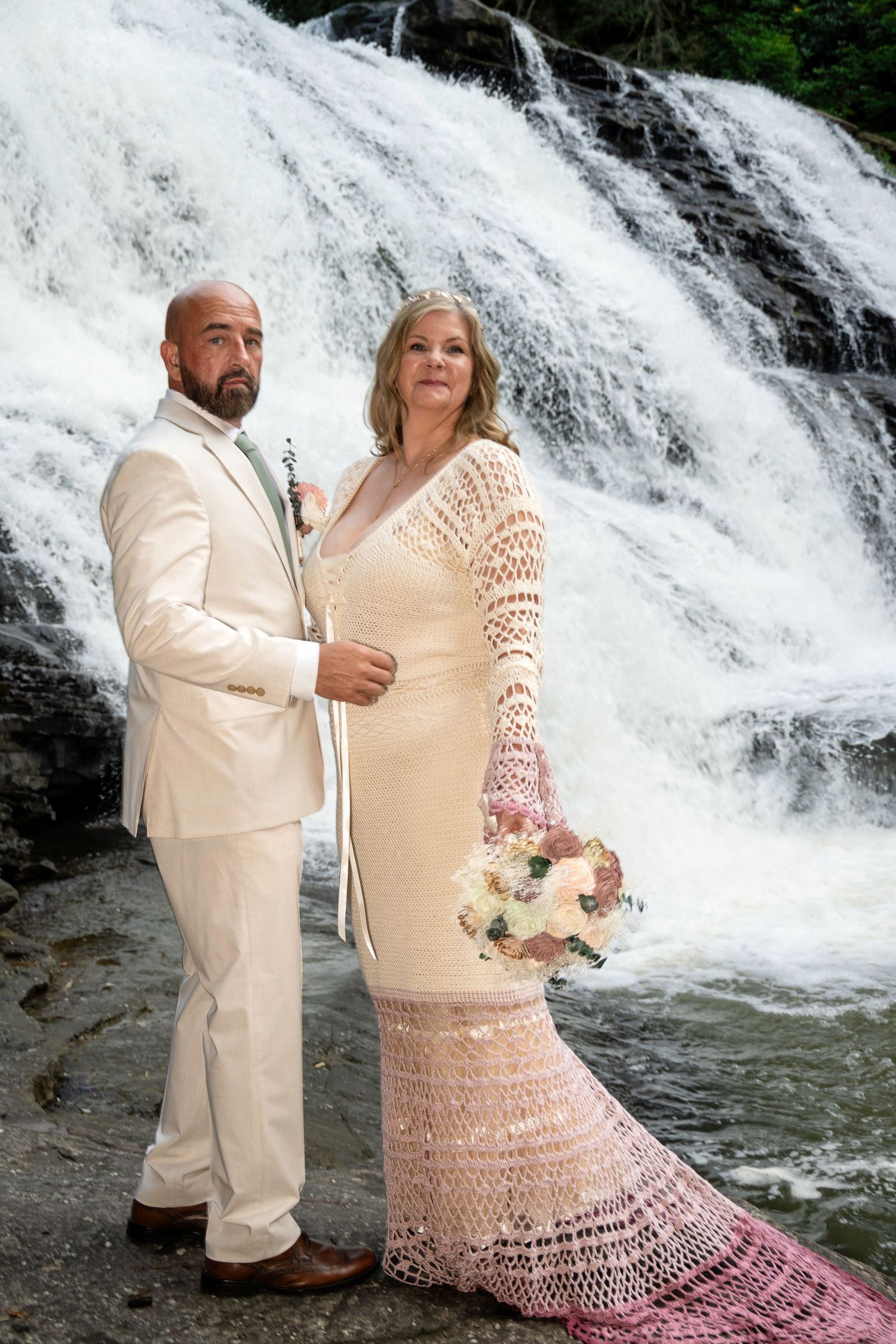 Couple in wedding attire poses before a waterfall, bride in crochet gown holding bouquet, groom in white suit.