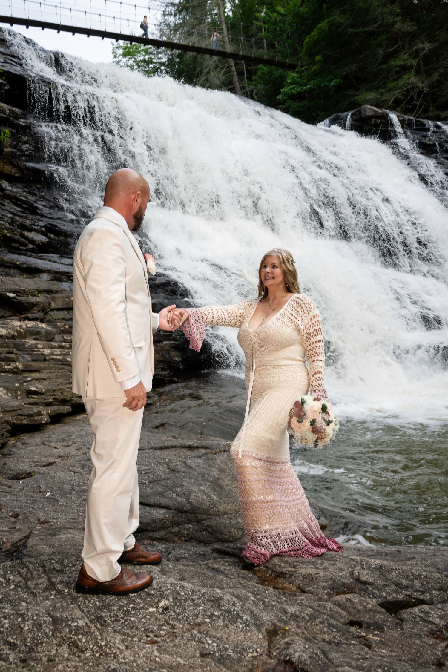Bride and groom hold hands before a cascading waterfall, she in lace gown with bouquet, he in cream suit.