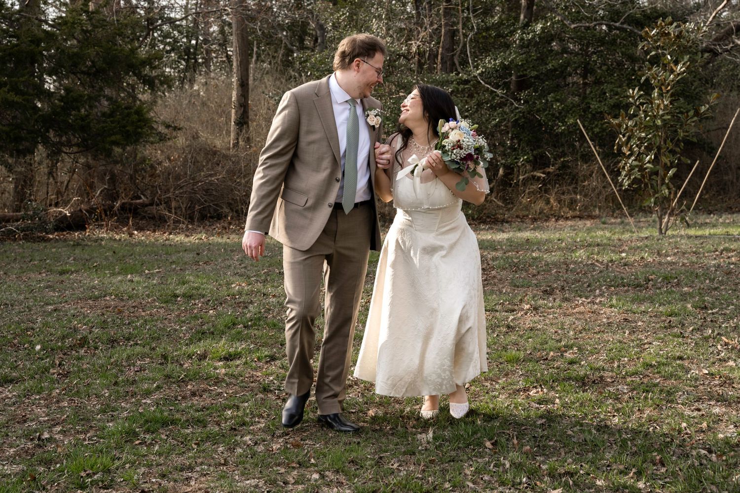 Bride and groom walking together outdoors on grass, smiling at each other, bride holding white bouquet, groom in tan suit.