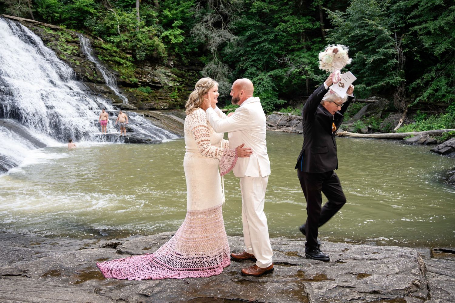 Bride and groom share a romantic moment on rocks before a stunning waterfall backdrop in a lush forest setting.