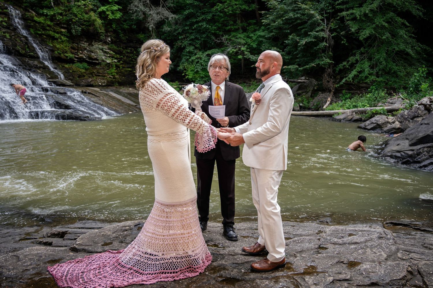 Couple exchanges vows before a waterfall with an officiant on rocky riverbank in a lush forest setting.