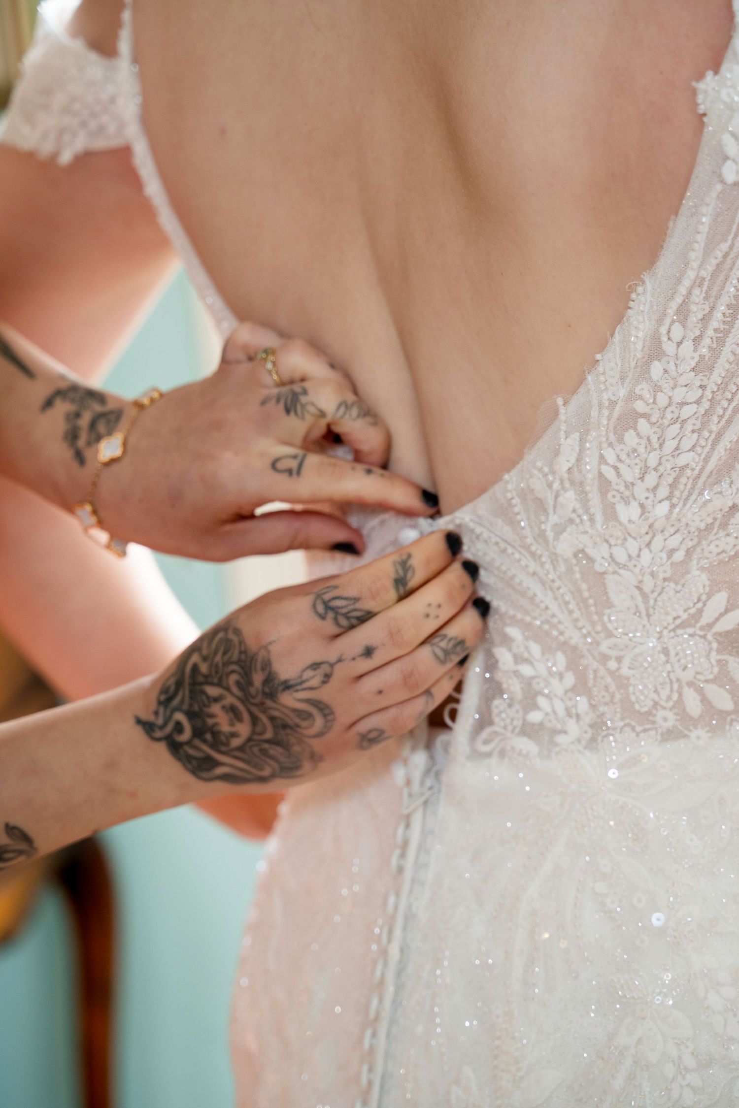 Tattooed hands fastening the back of a white lace wedding dress on a bride.