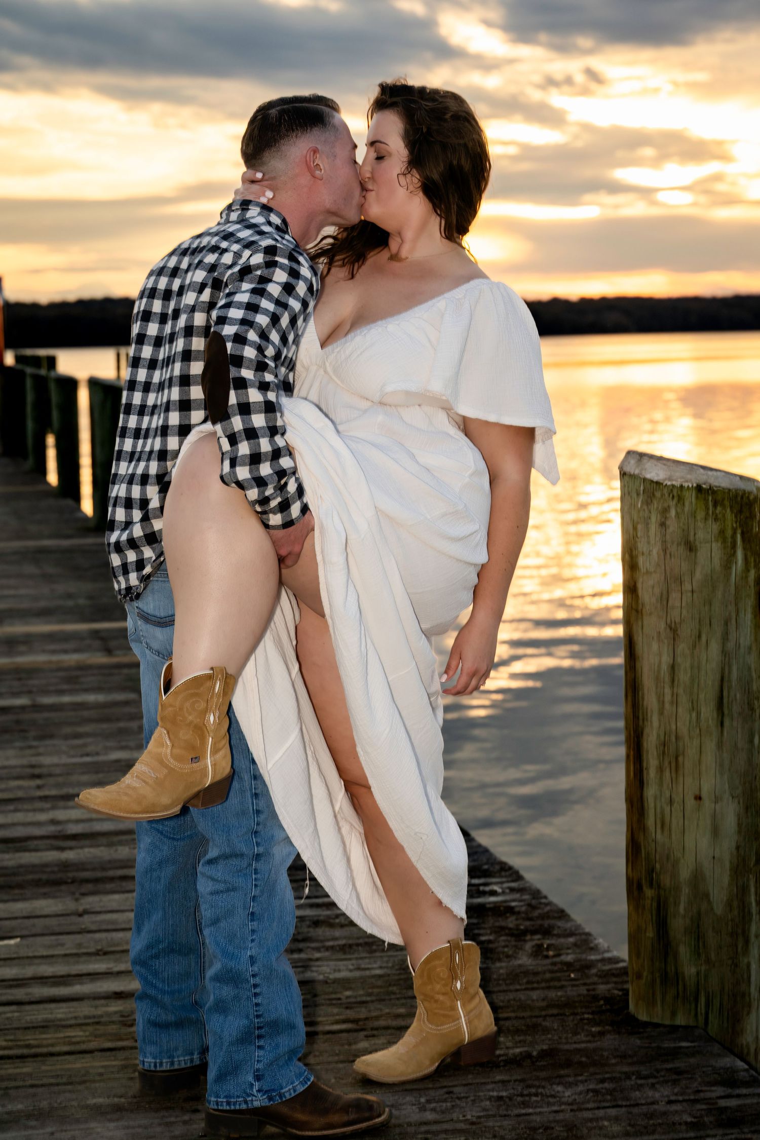 Couple sharing a passionate kiss on a wooden dock at golden sunset by the water.